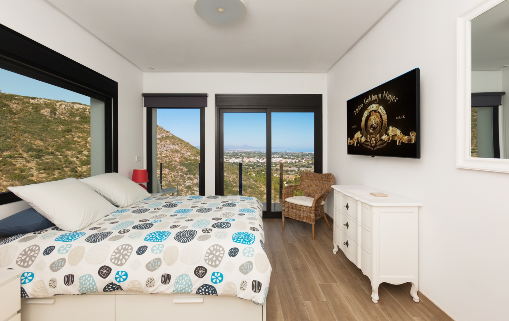 Bedroom featuring panoramic mountain and sea views through large black-framed windows, wood-effect flooring, a white dresser, and a wall-mounted television.