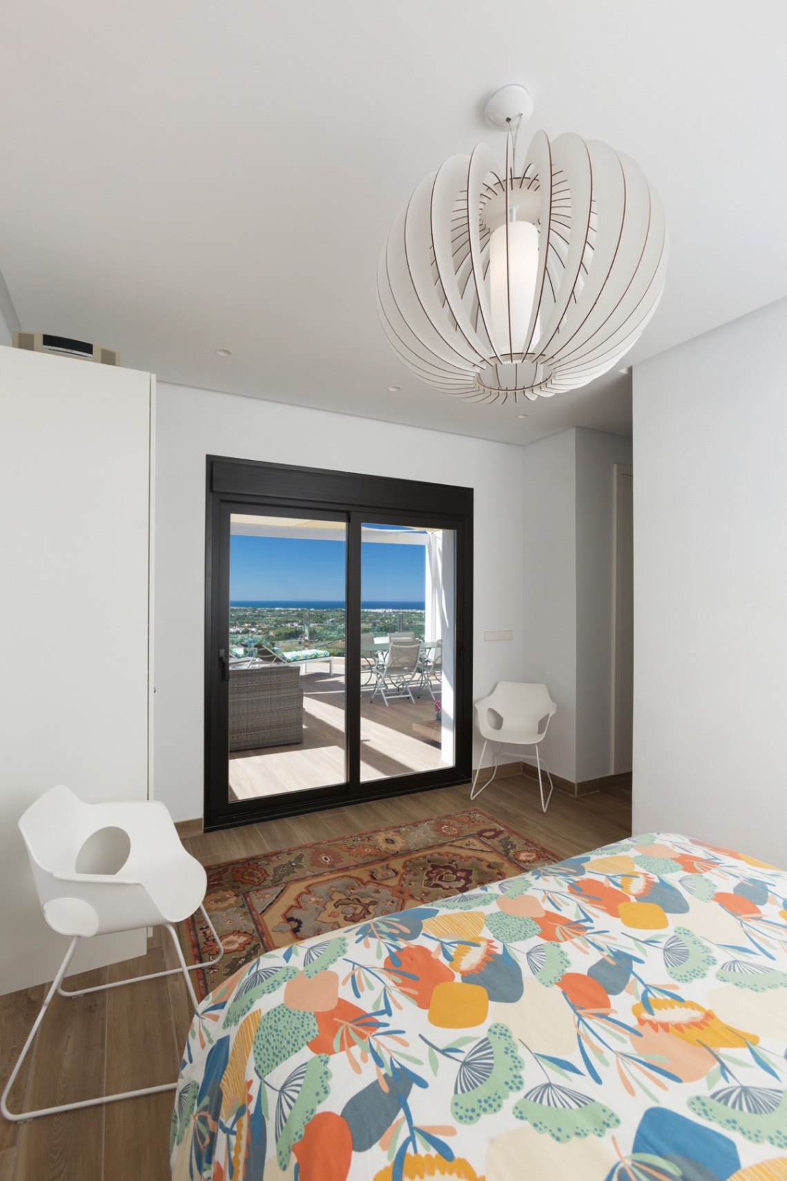 Bright bedroom with direct terrace access through black-framed sliding doors, featuring a designer pendant light and panoramic sea views.