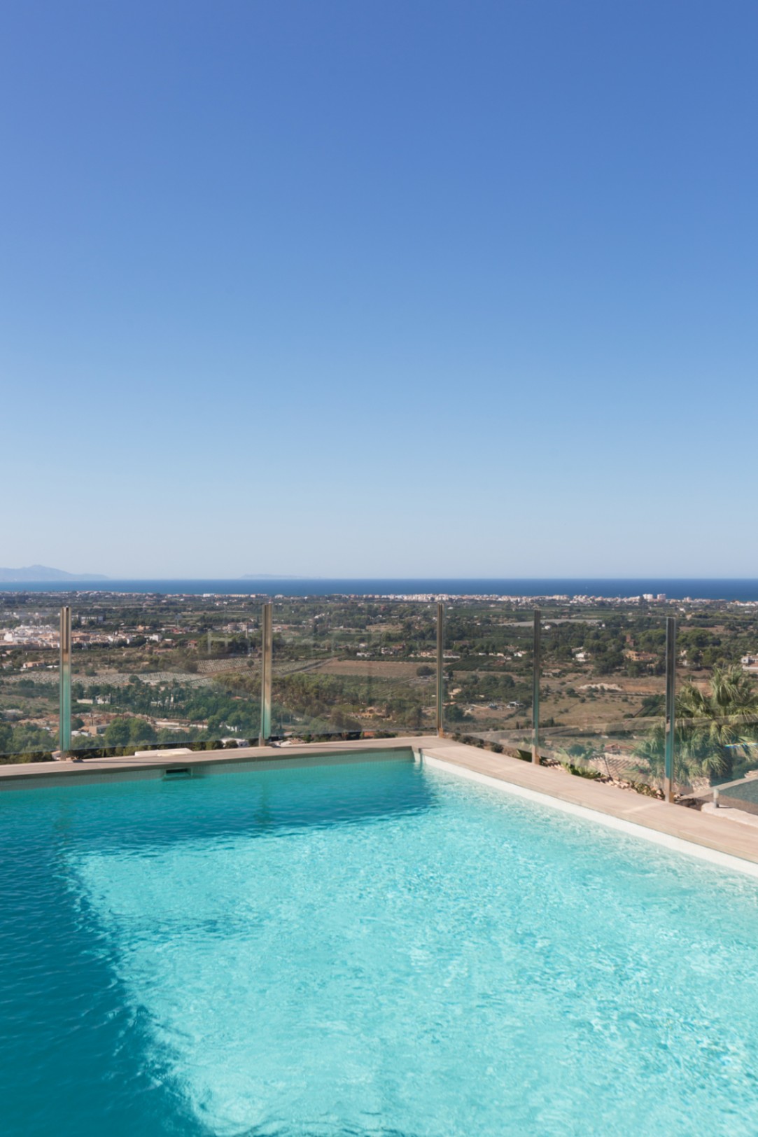 Elevated infinity pool with a glass safety railing, offering panoramic views of the coastal valley and the Mediterranean Sea under a clear sky.