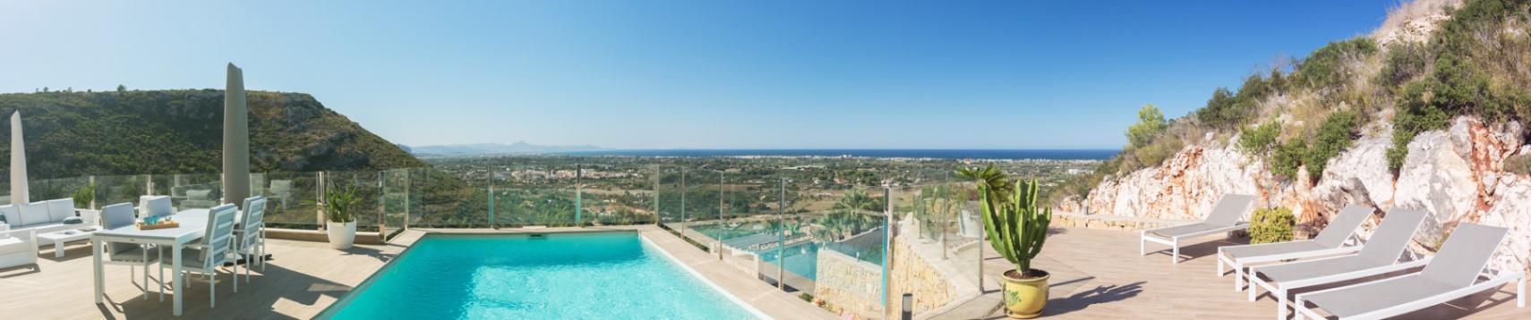 Elevated terrace featuring an infinity pool, glass railings, and white outdoor furniture with panoramic views of the Mediterranean coastline.
