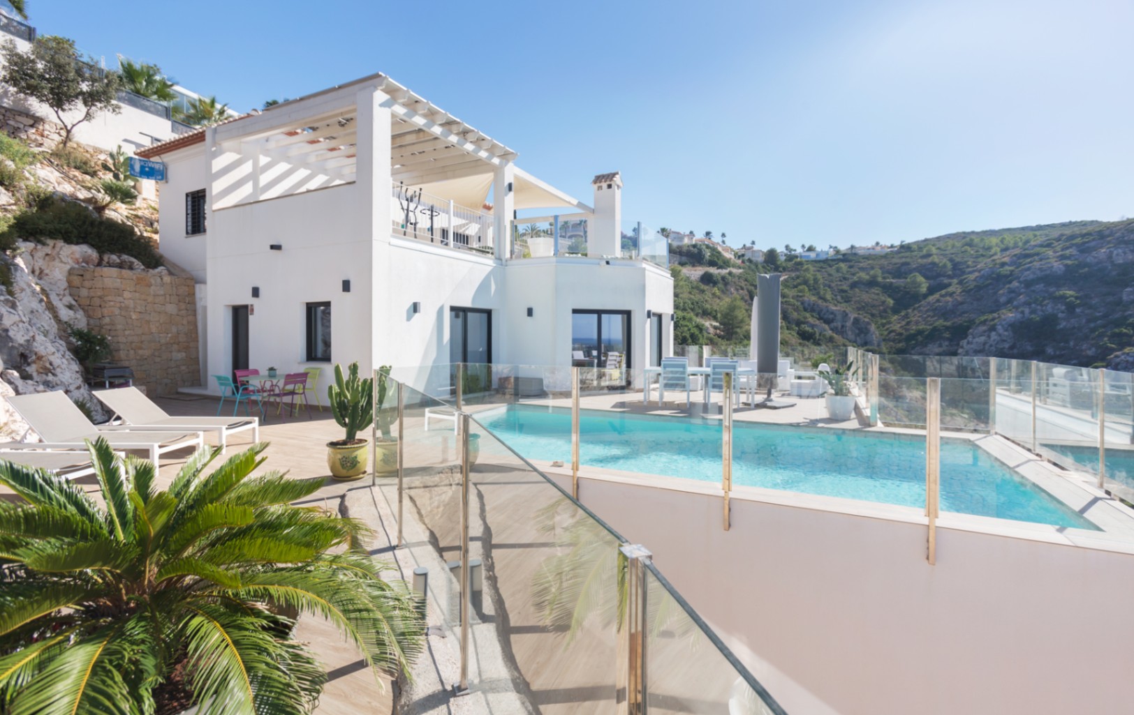 Exterior view of a white multi-level villa featuring an infinity pool with glass railings, sun terraces, and panoramic hillside views.