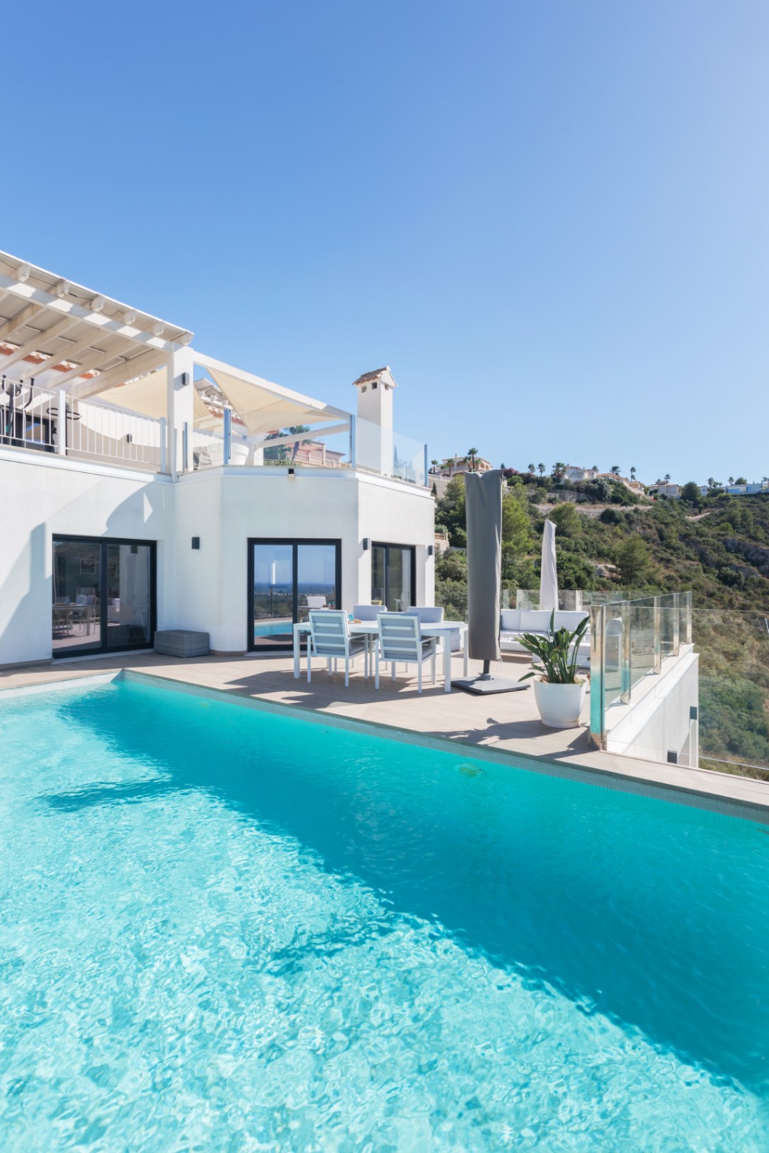 Infinity pool with turquoise water bordering a tiled terrace featuring a white dining set and glass balustrades overlooking the hillside.