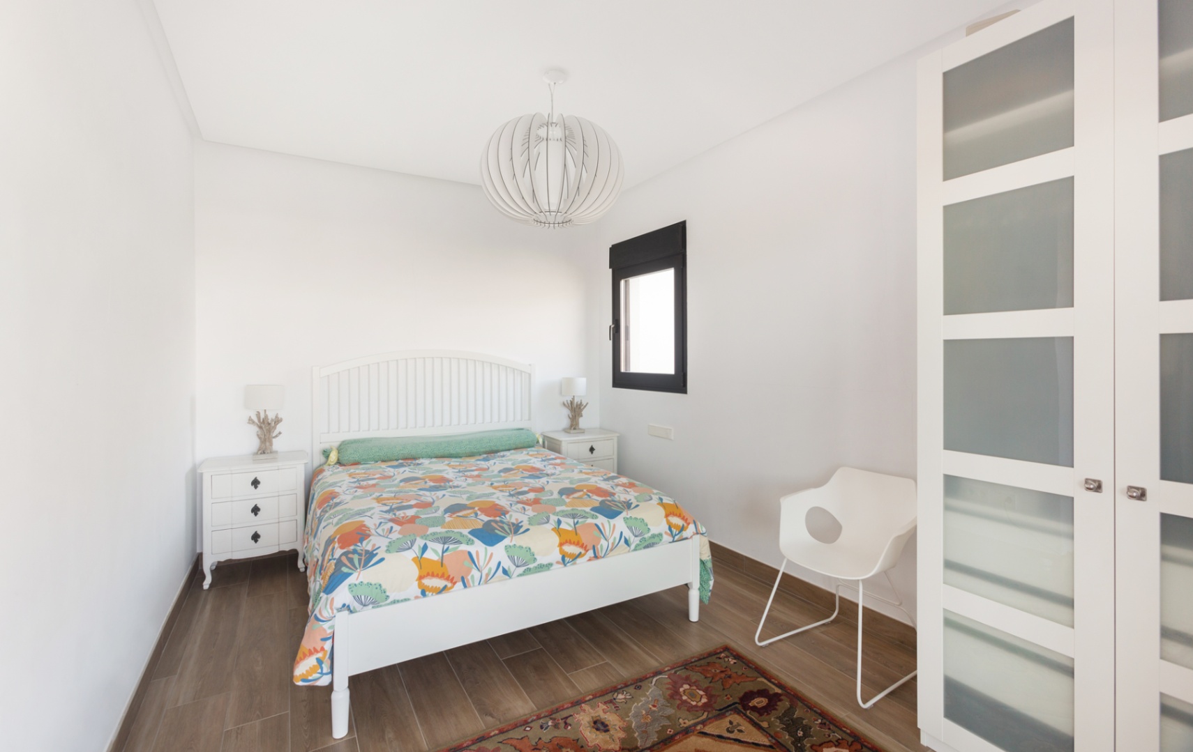 Luminous bedroom featuring a white wooden bed frame, matching nightstands, and a modern wardrobe with frosted glass. Wood-effect flooring and a designer pendant light complete the space.