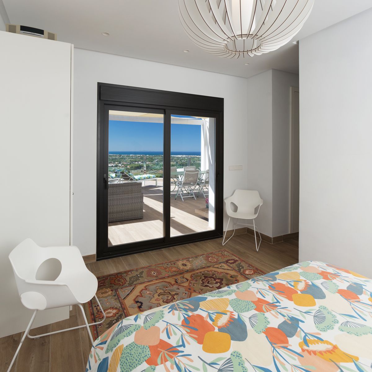 Modern bedroom featuring a large sliding glass door that opens onto a furnished terrace with panoramic sea views. Includes a sculptural pendant light and white designer chairs.