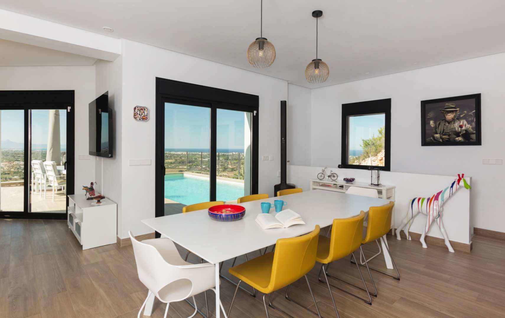 Modern dining area featuring a white table with yellow chairs, wood-effect flooring, and sliding doors opening to a pool terrace with panoramic coastal views.