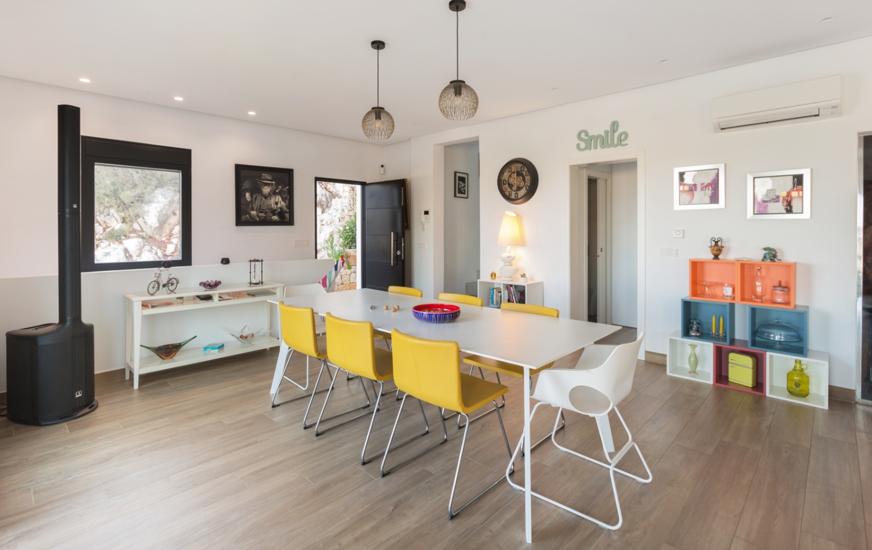 Modern dining space featuring a white table with yellow chairs, wood-effect flooring, a sleek black wood-burning stove, and colorful modular shelving.