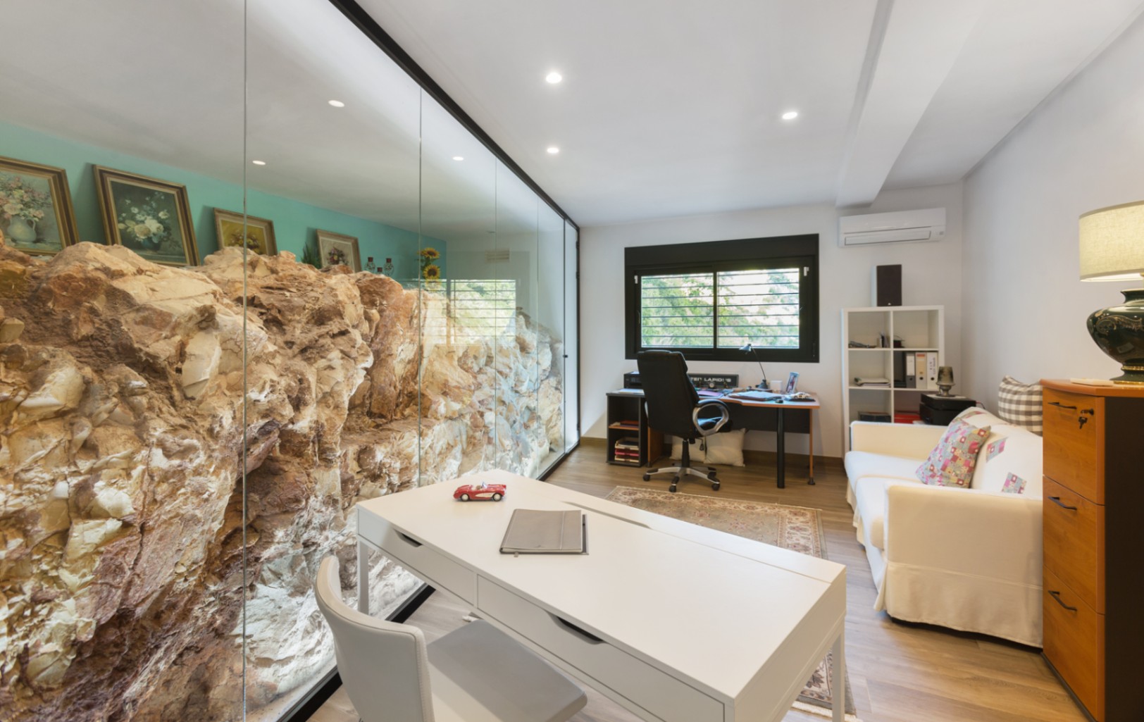 Modern home office featuring a floor-to-ceiling glass wall showcasing a natural rock formation. Includes a white desk, sofa, and recessed ceiling lighting.