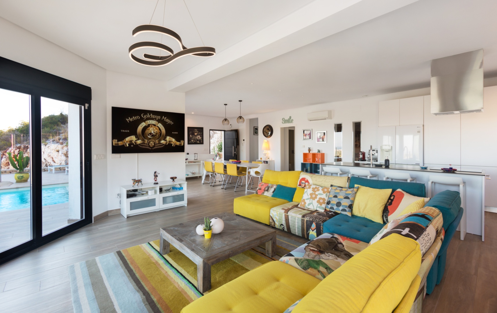 Open-plan living space featuring a colorful modular sofa, wooden coffee table, and seamless access to a terrace with a private pool through large sliding glass doors.