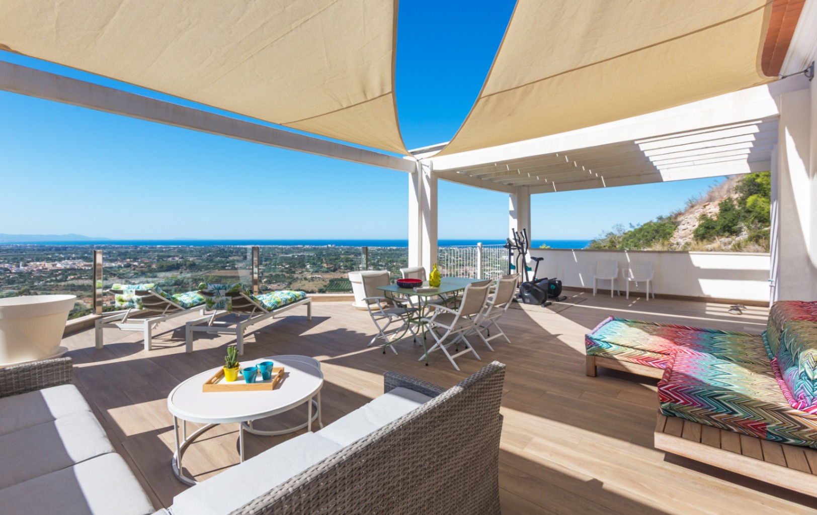 Spacious rooftop terrace with wood-effect flooring, sail shades, and panoramic sea views. Includes a dining area, lounge seating, and sunbeds under a white pergola structure.