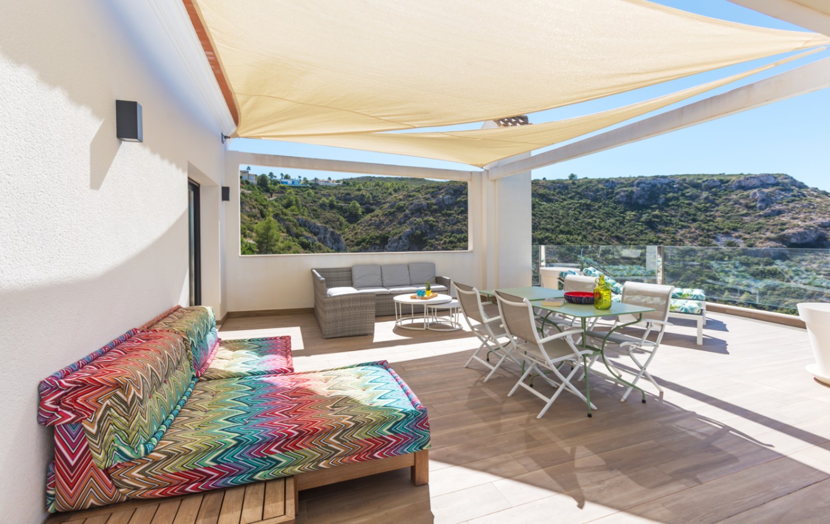 Spacious terrace with beige shade sails, featuring a dining set, wicker lounge, and colorful daybed overlooking a green valley through glass railings.