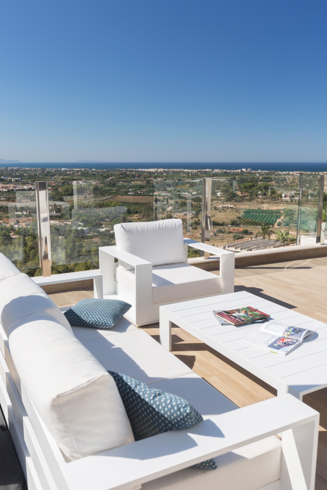 Terrace with modern white lounge furniture and glass railings, offering panoramic views of the coastal landscape and the Mediterranean Sea.