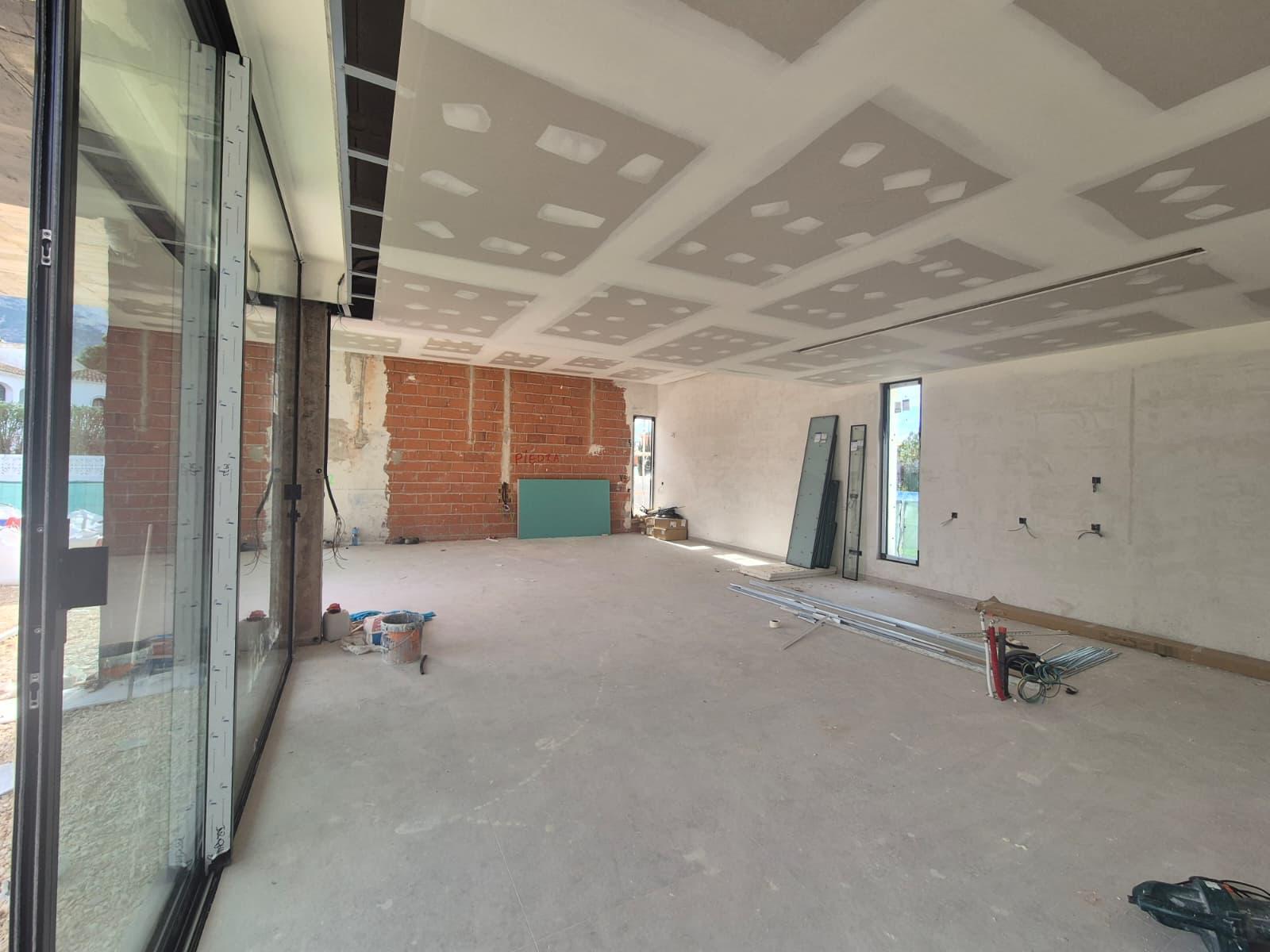Spacious living area under construction featuring a plasterboard ceiling with lighting cutouts, exposed brickwork, and large floor-to-ceiling glass sliding doors.