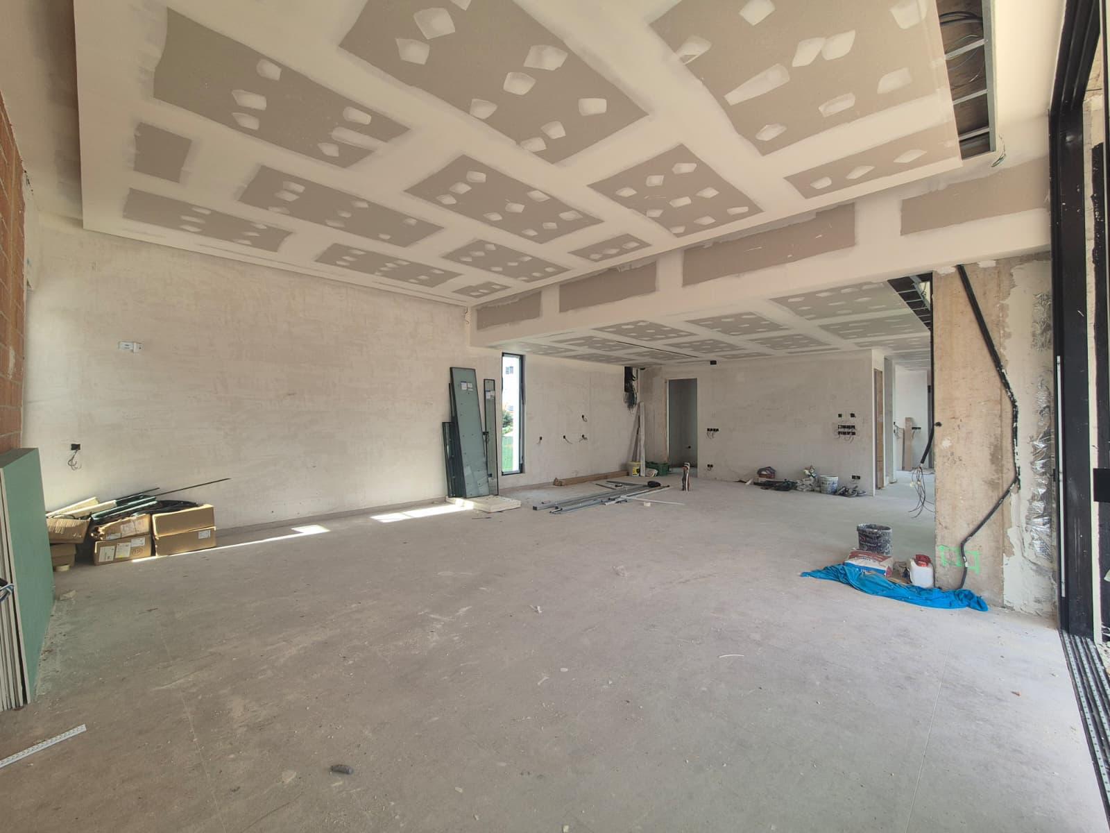 Spacious open-plan interior under construction featuring multi-level plasterboard ceilings with recessed lighting trays and large floor-to-ceiling window openings.
