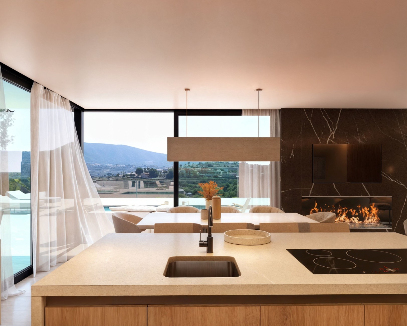 Modern open-plan kitchen featuring a stone island with integrated cooktop, adjacent to a dining area and a living space with a dark marble fireplace and mountain views.