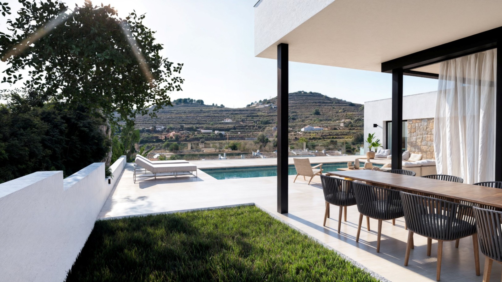 Modern outdoor terrace with a wooden dining set, infinity pool, and sun loungers, overlooking a terraced landscape and hills under bright daylight.