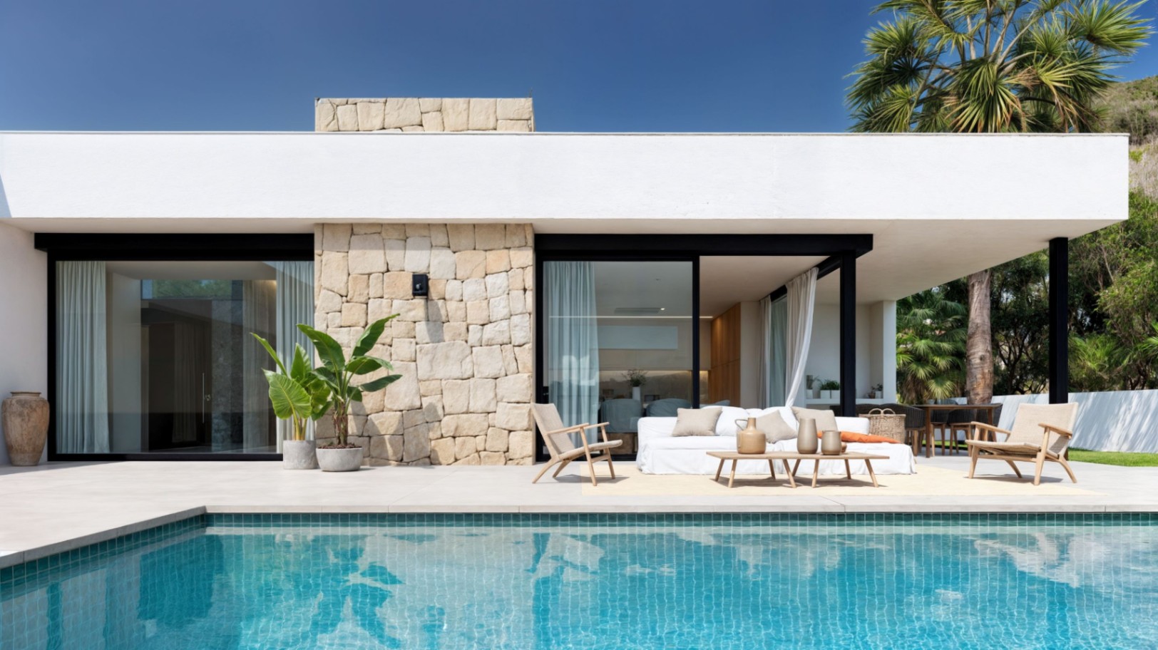 Modern villa exterior featuring a turquoise tiled pool, dry stone accent walls, and a lounge area on a spacious tiled terrace under a flat roof.