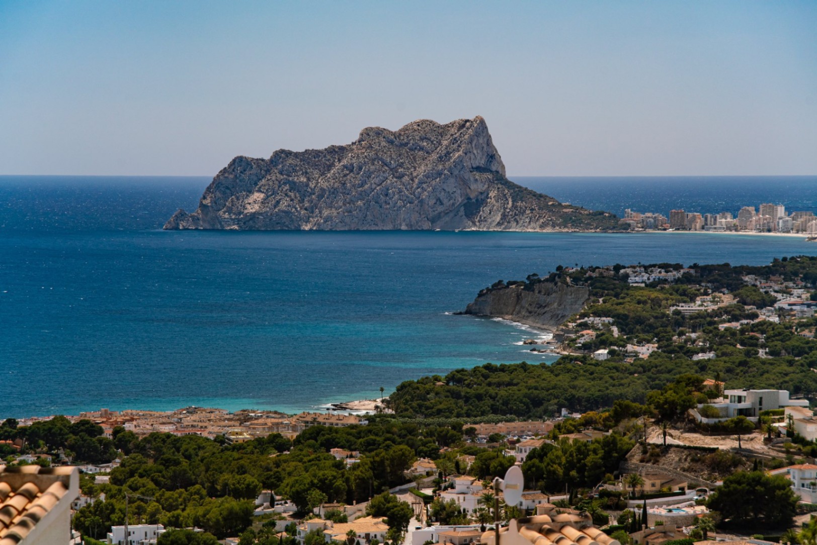 Elevated panoramic view of the Mediterranean coastline featuring the Peñón de Ifach, turquoise waters, and a residential area nestled among dense pine forests.