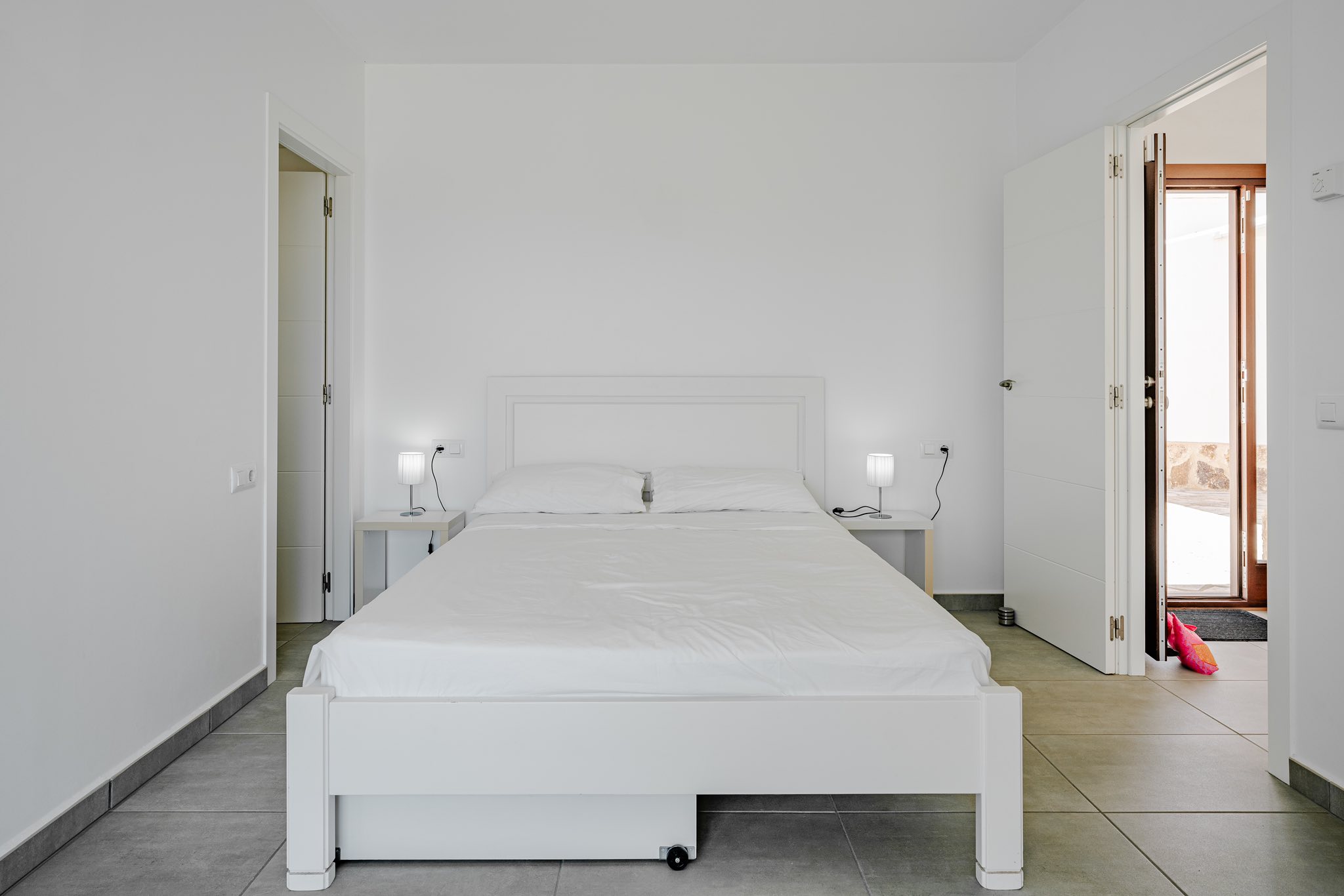 Minimalist bedroom featuring a white wooden bed frame, matching nightstands with lamps, and grey tiled flooring. Open doors lead to adjacent spaces and an outdoor area.