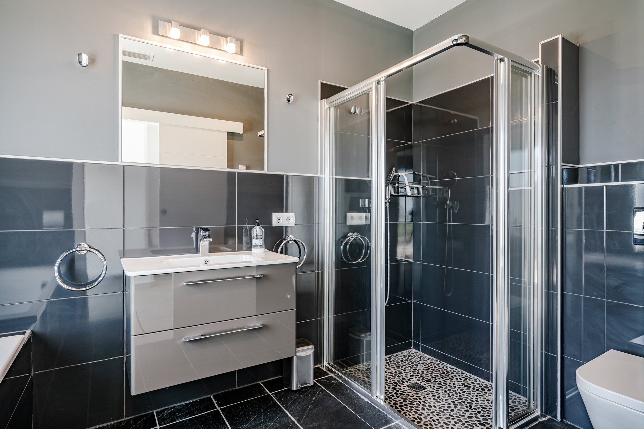 Modern bathroom featuring charcoal glossy tiles, a floating vanity with integrated sink, and a glass-enclosed shower with a pebble-patterned floor.