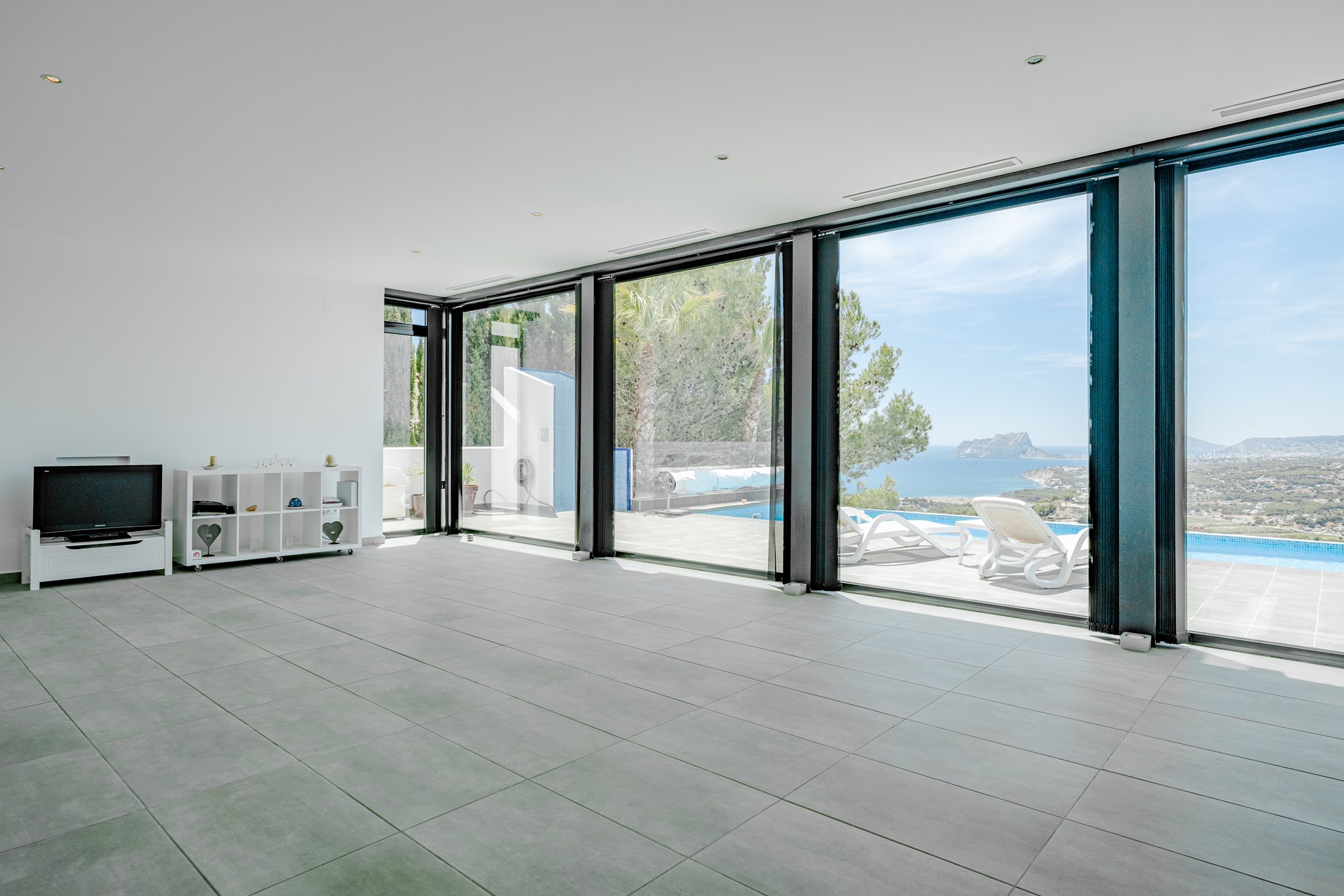 Spacious living area featuring grey tiled flooring and floor-to-ceiling glass doors opening to a pool terrace with views of the Mediterranean and Peñón de Ifach.
