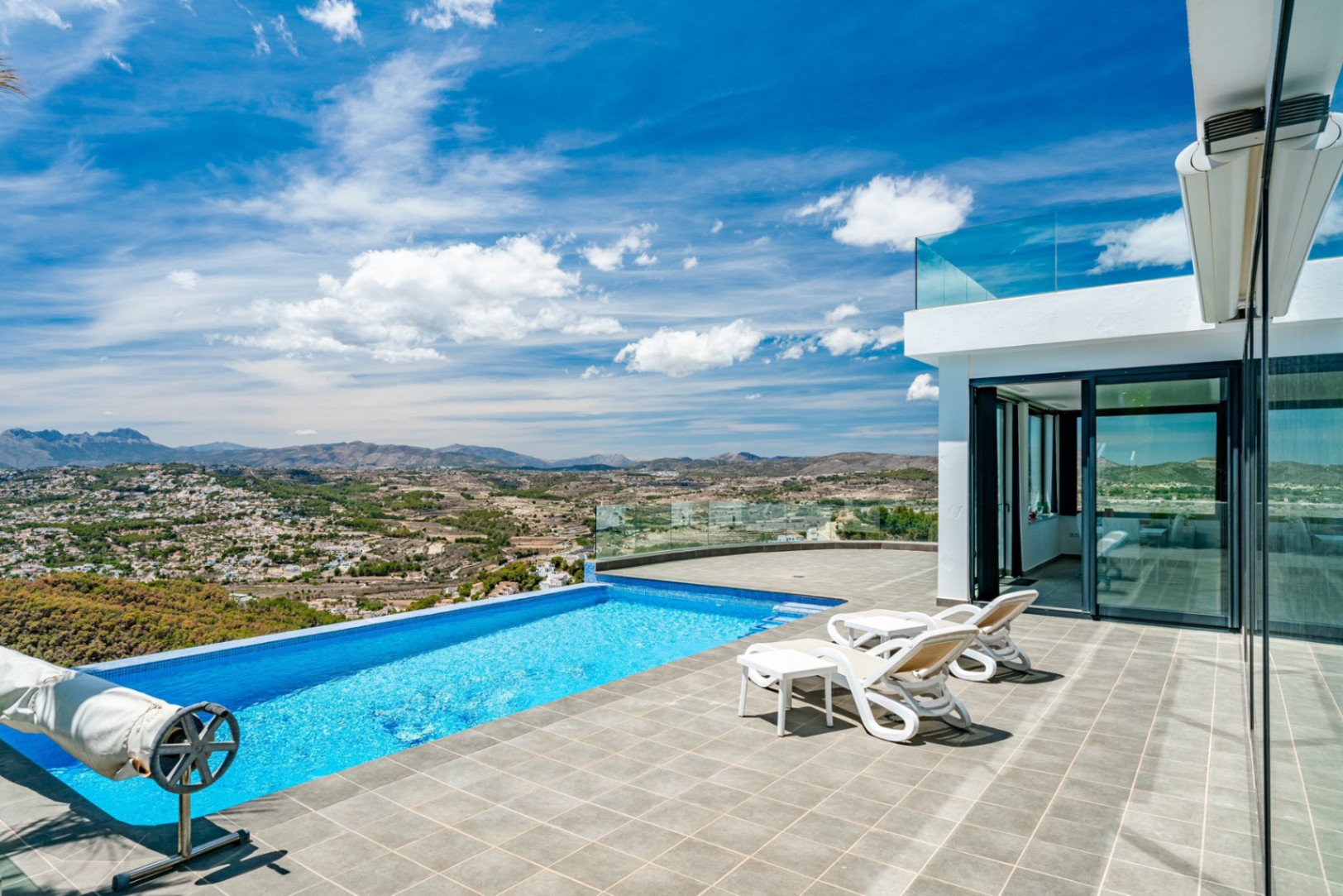 Terrace with an infinity pool and glass railings, offering panoramic valley and mountain views. Features stone tiling, sun loungers, and modern white architecture.