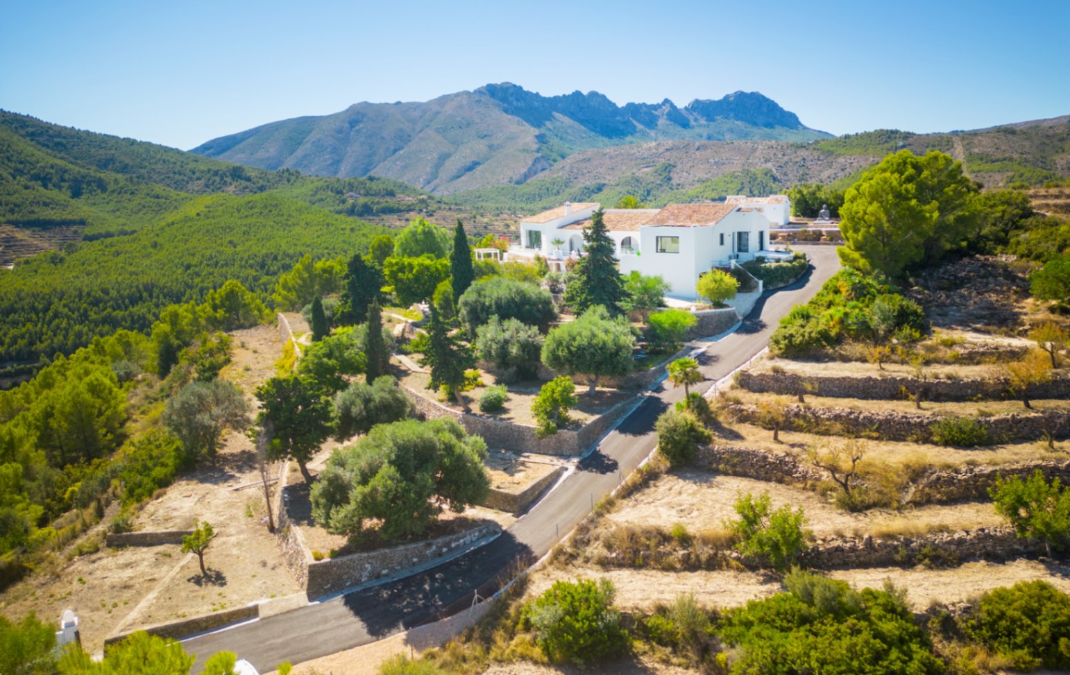 Aerial view of a white villa nestled in a mountainous landscape with terraced stone walls, Mediterranean gardens, and panoramic views of the pine forest.