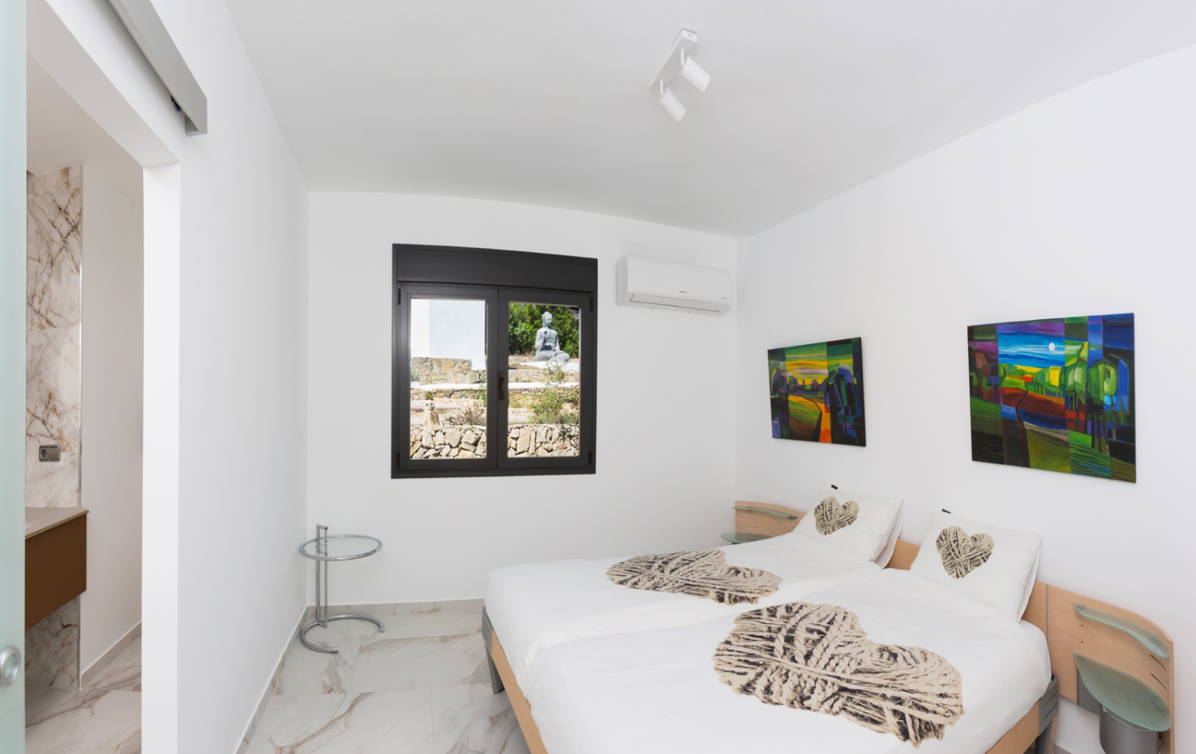 Bedroom featuring twin beds, marble-effect tiling, and a black-framed window overlooking a stone garden. Includes air conditioning and ensuite bathroom access.