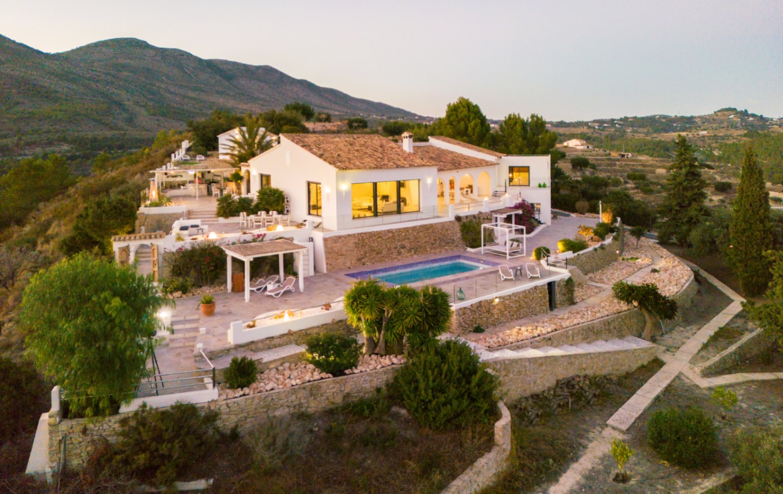 Elevated view of a Mediterranean villa featuring white facades, terracotta roofing, and multi-level terraces with a swimming pool and stone retaining walls.