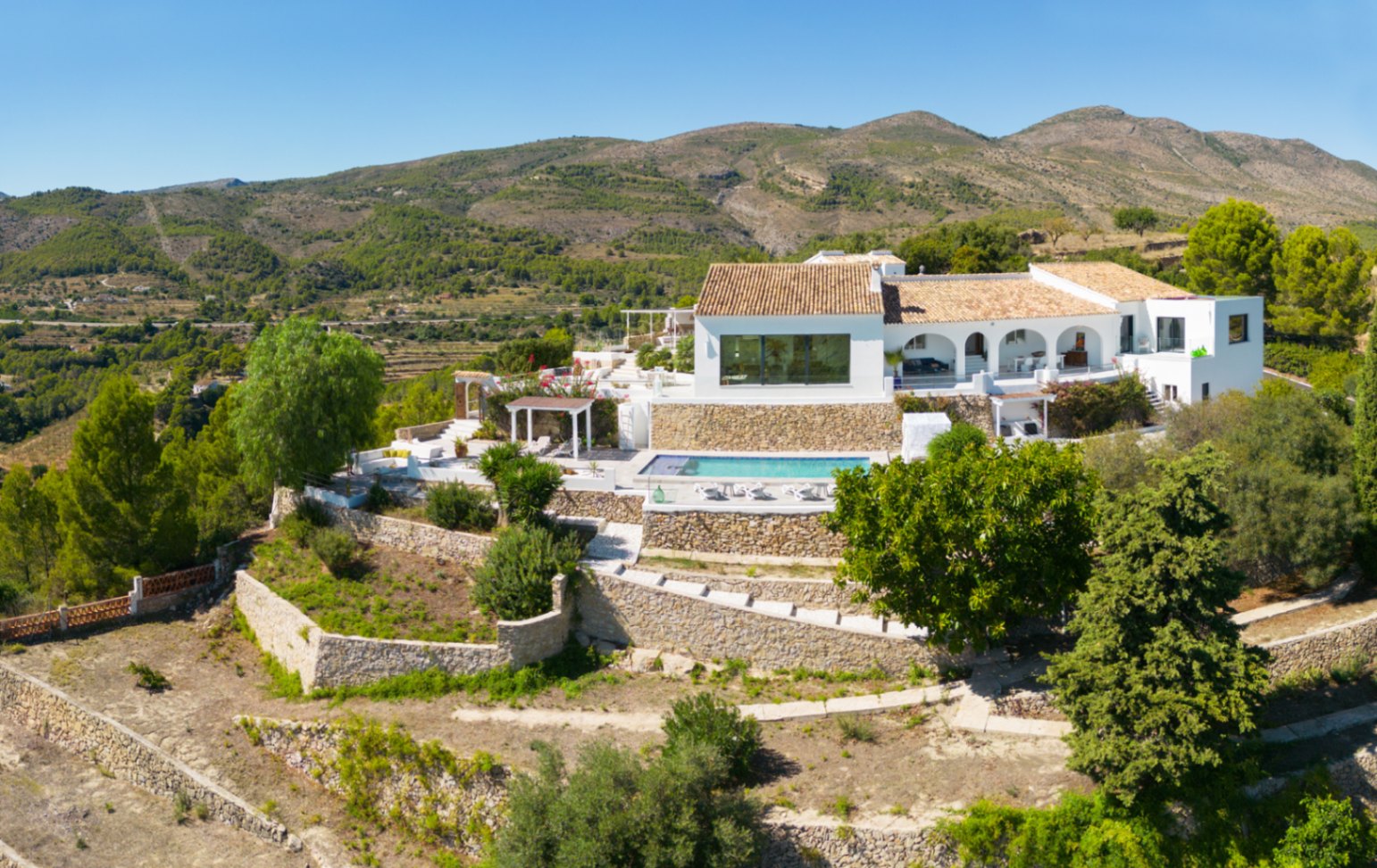 Elevated view of a white Mediterranean villa featuring stone-terraced gardens, a private swimming pool, and an arched porch overlooking the mountain landscape.