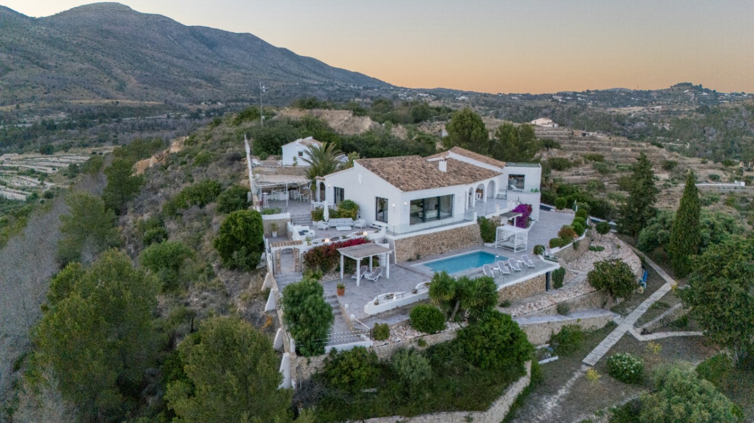 Elevated view of a white Mediterranean villa featuring terraced stone walls, a swimming pool, and multiple outdoor lounge areas set against a mountain backdrop.