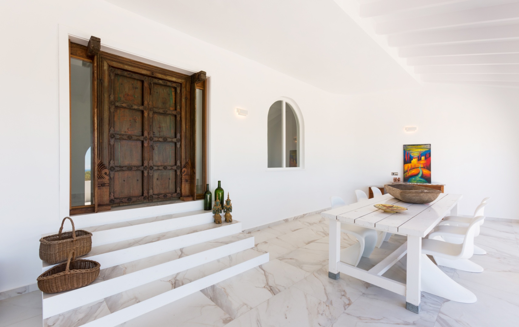 Entrance featuring an antique carved wooden door, white marble flooring, and an outdoor dining area with a rustic table and modern Panton-style chairs.