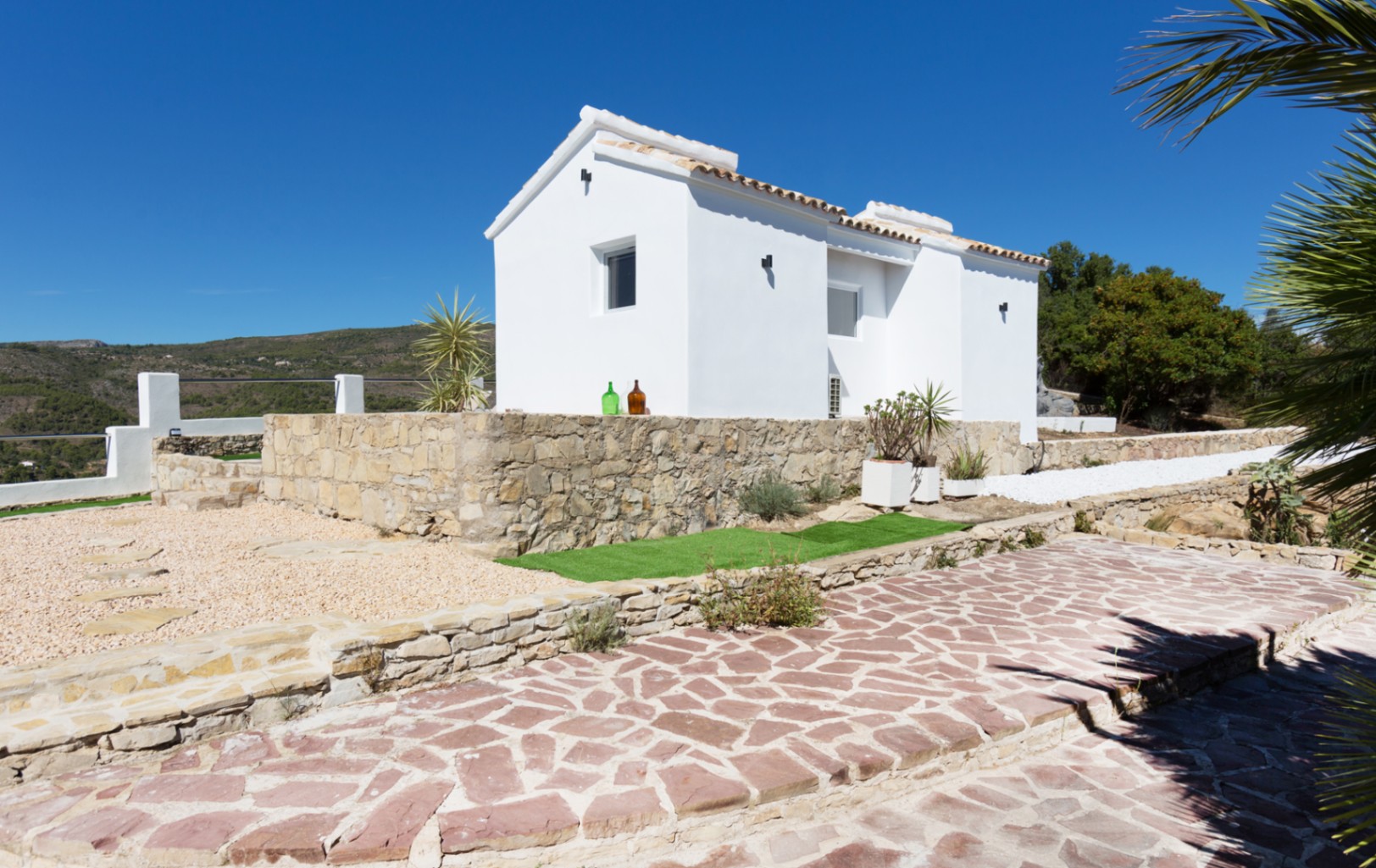 Exterior view of a white-washed villa featuring a traditional stone retaining wall, terracotta tile roof, and a paved stone pathway under a clear blue sky.