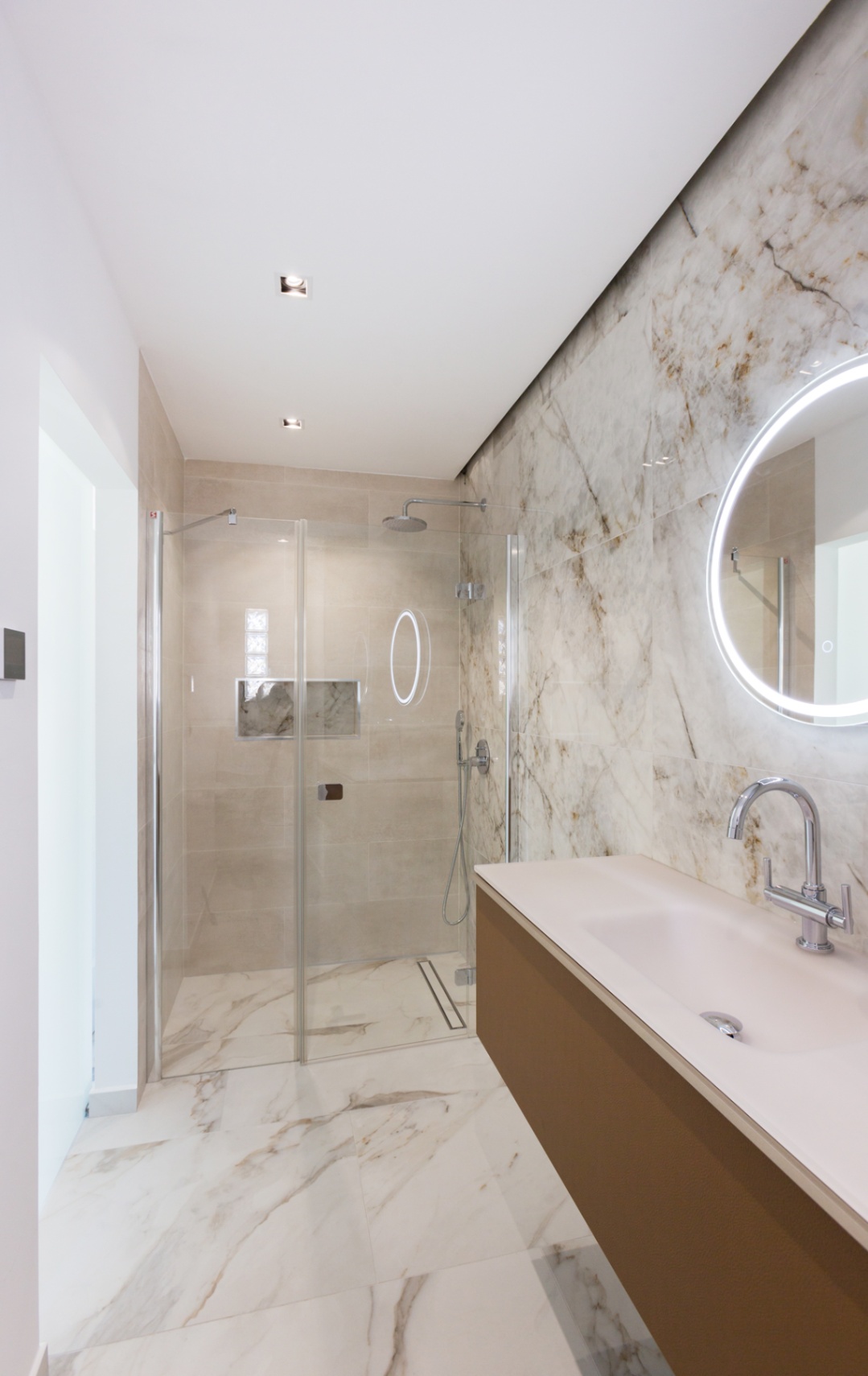 Modern bathroom featuring a walk-in glass shower, marble-effect wall tiling, and a floating vanity with an integrated sink and circular backlit LED mirror.