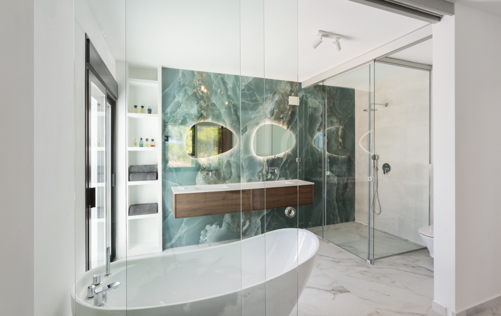 Modern bathroom with a green onyx feature wall, freestanding bathtub, and floating wood vanity. Features organic backlit mirrors and a glass-enclosed walk-in shower.