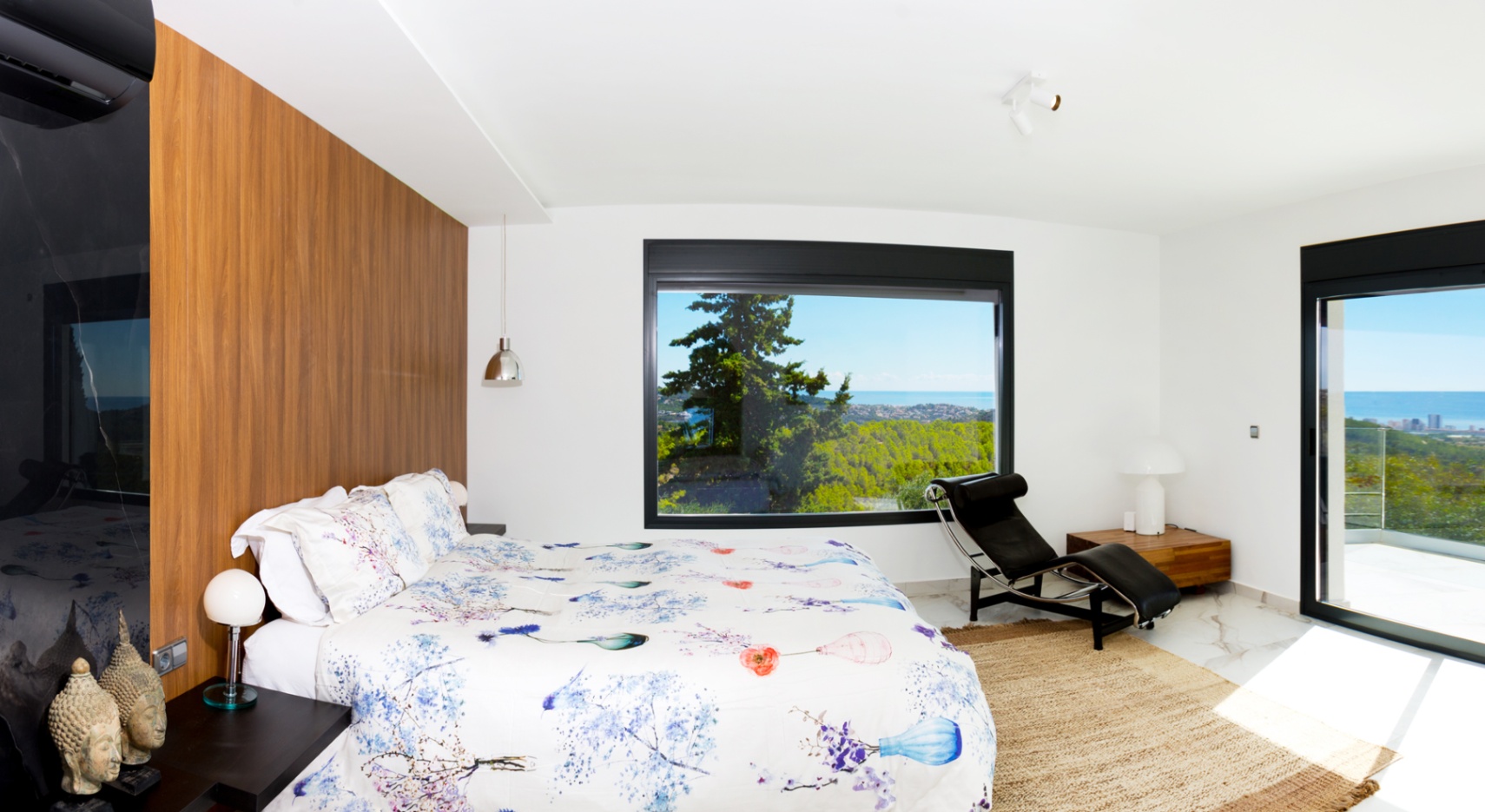 Modern bedroom featuring a wood-paneled accent wall, marble flooring, and a large picture window offering panoramic sea and forest views. Includes a designer chaise longue.