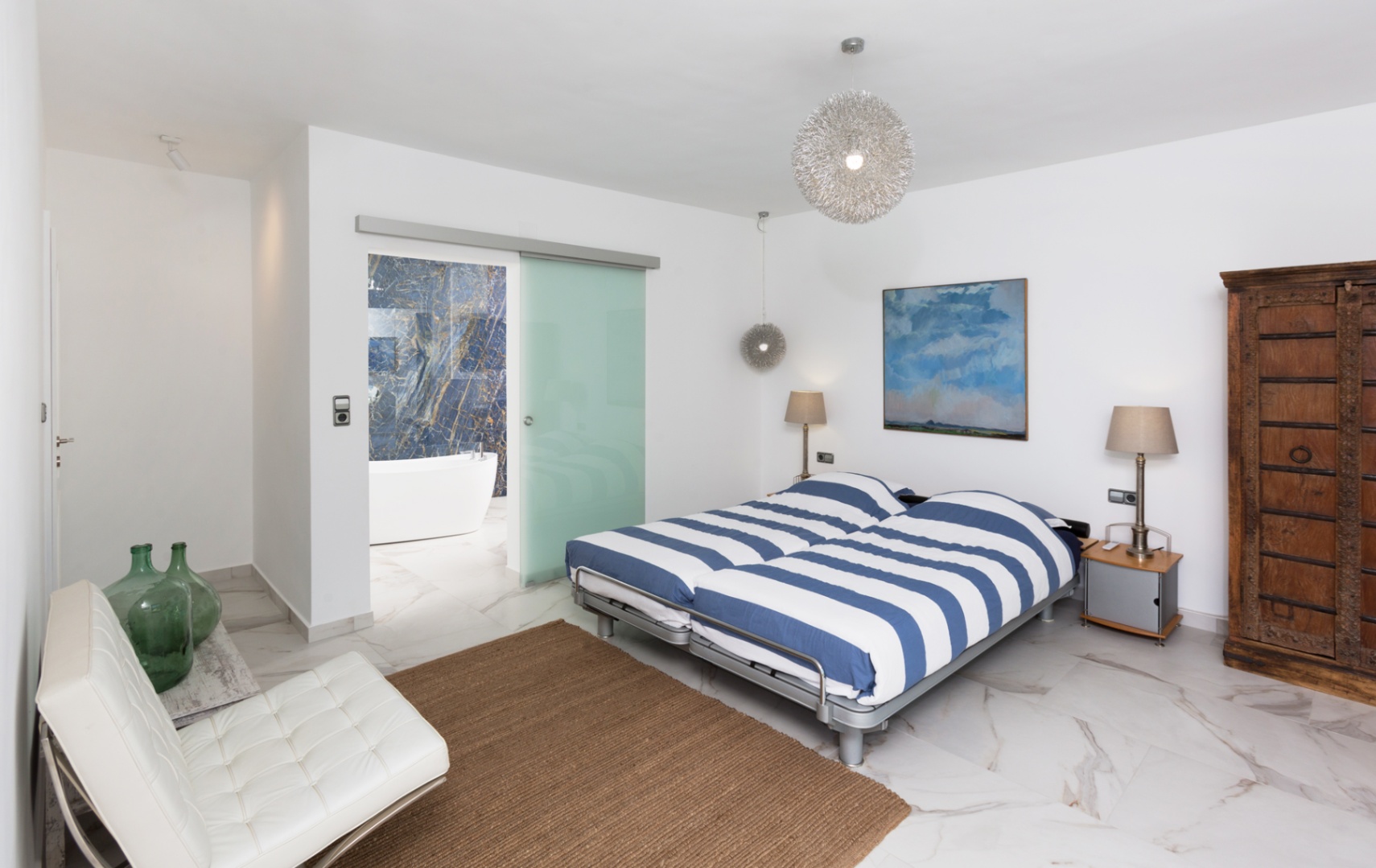 Modern bedroom with marble-effect flooring, blue-striped bedding, and a frosted glass door opening to an en-suite bathroom with a blue stone wall and freestanding tub.