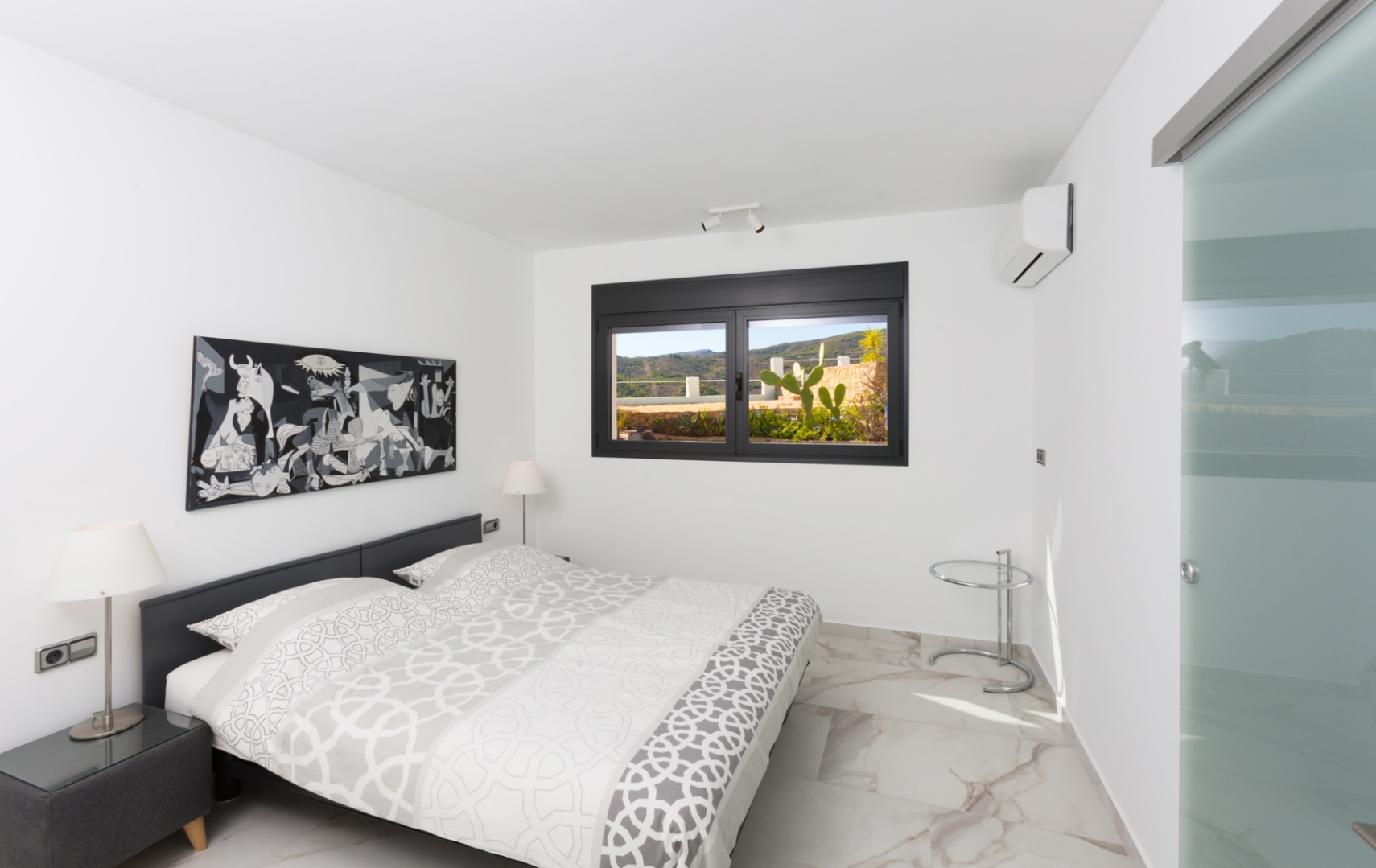 Modern bedroom with marble-effect porcelain flooring, a monochrome art piece, and a black-framed window overlooking a terrace with cacti and hills.