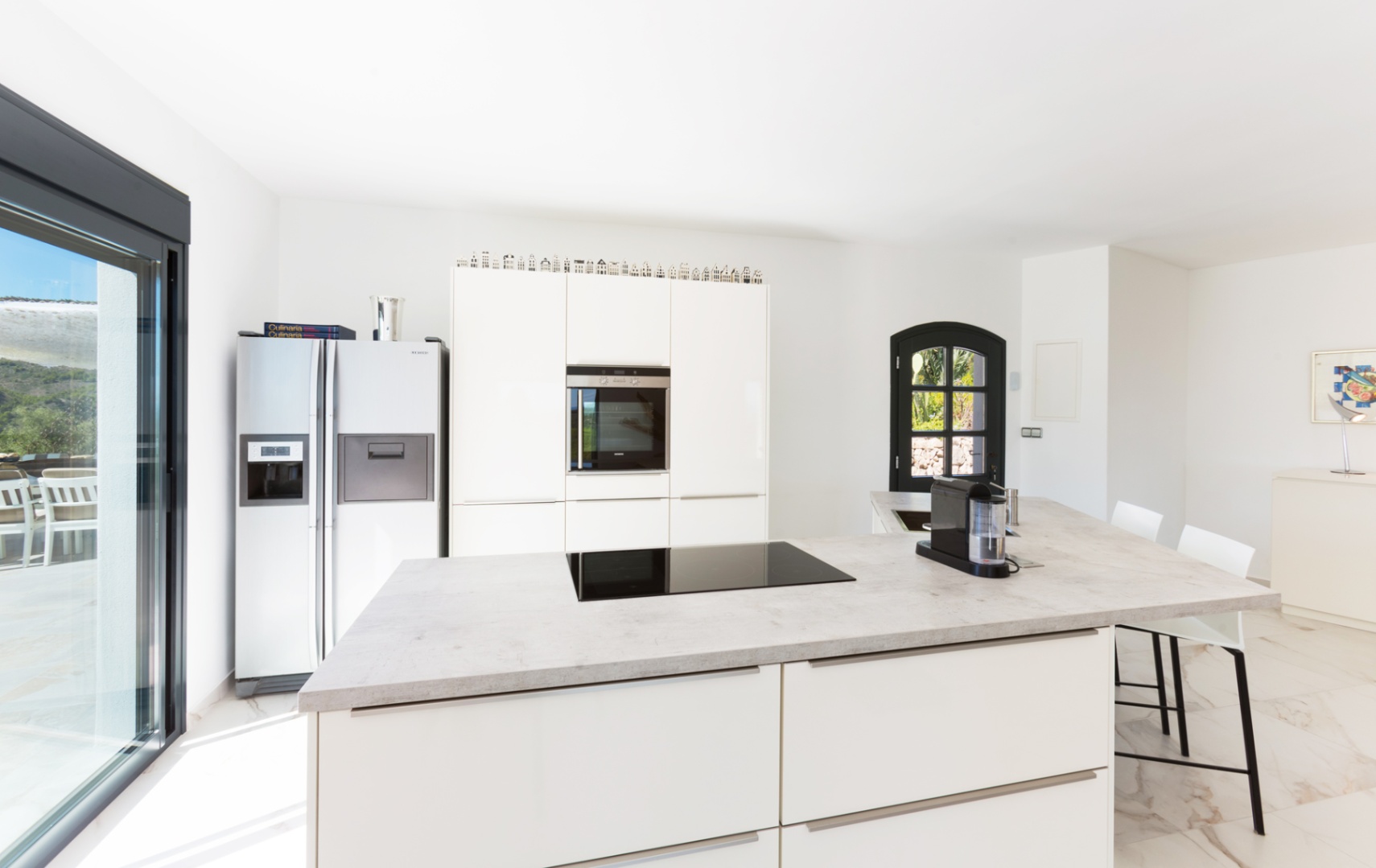 Modern kitchen featuring a large island with a stone-effect countertop, integrated induction hob, white cabinetry, and a double-door refrigerator.