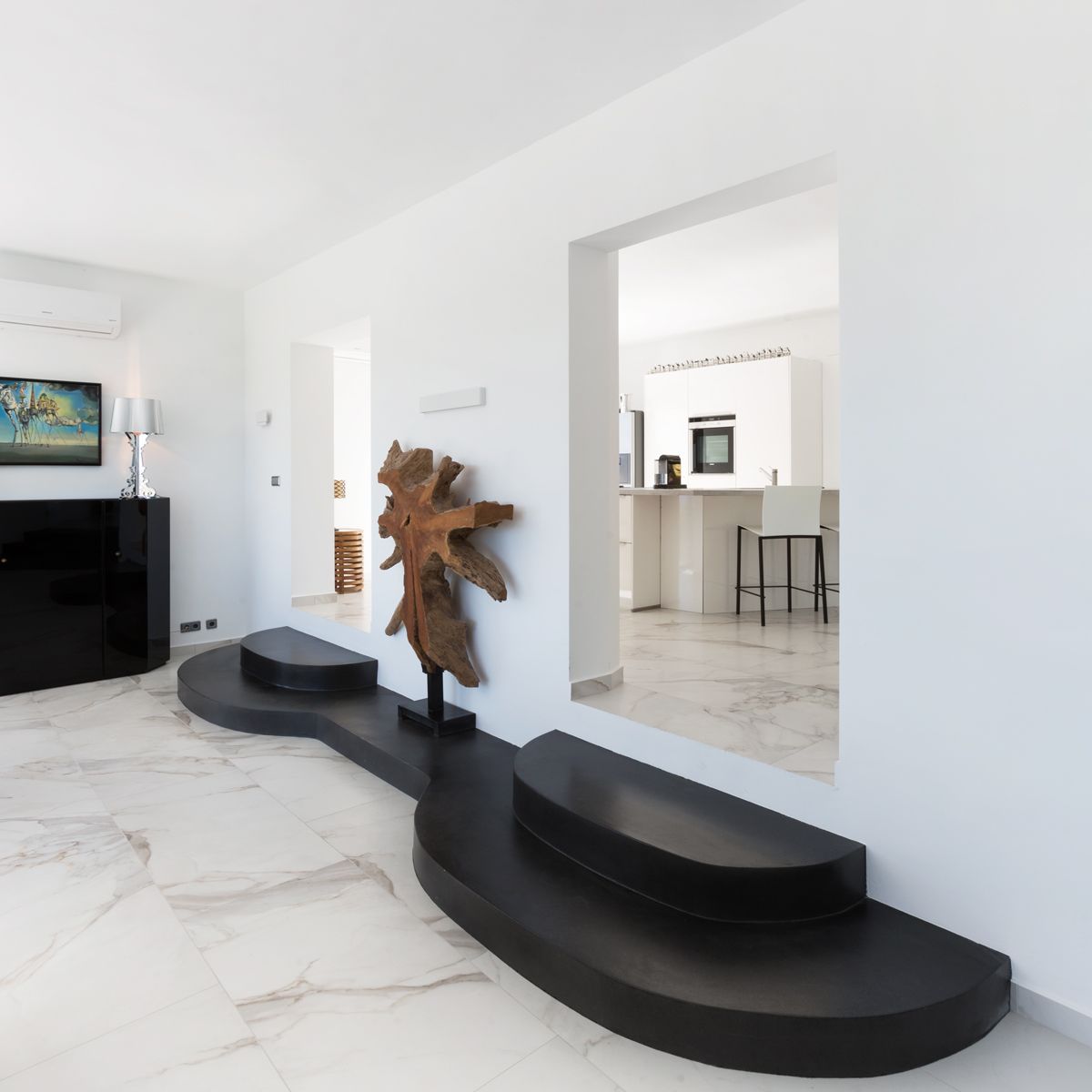 Modern living area featuring white marble floors and a unique black curved platform. A large natural wood sculpture sits between openings to a minimalist white kitchen.