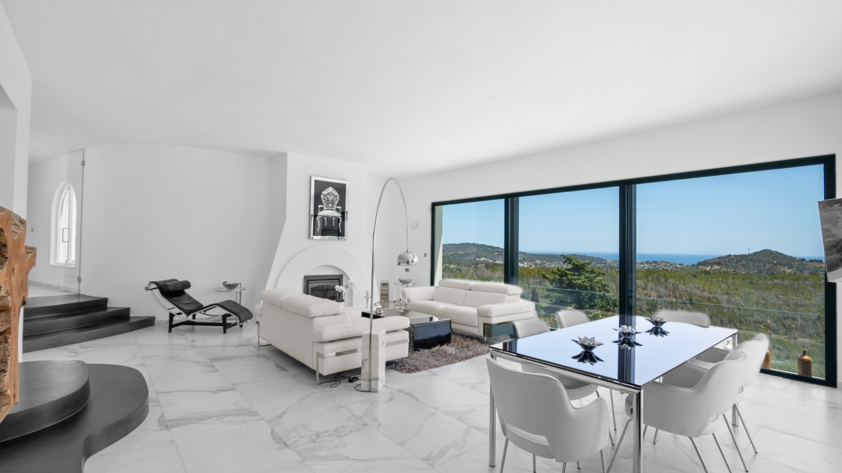 Modern open-plan living area featuring white marble floors, a built-in fireplace, and floor-to-ceiling windows with panoramic views of the Mediterranean landscape.