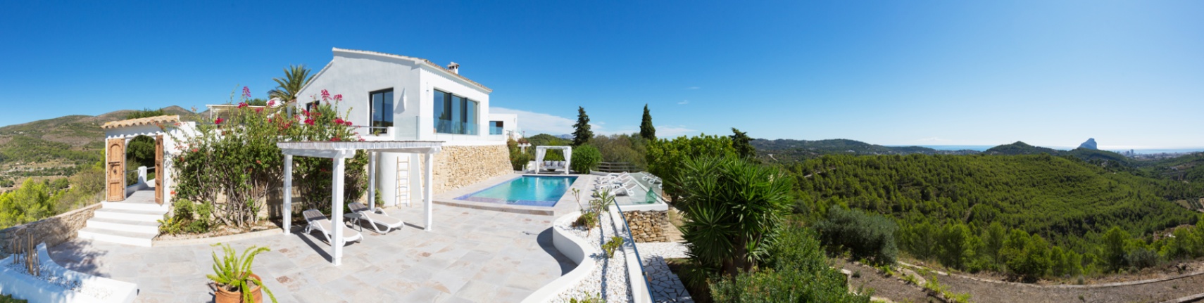 Panoramic terrace featuring a swimming pool, white pergola, and stone paving, overlooking a lush valley with distant views of the Mediterranean Sea and Peñón de Ifach.