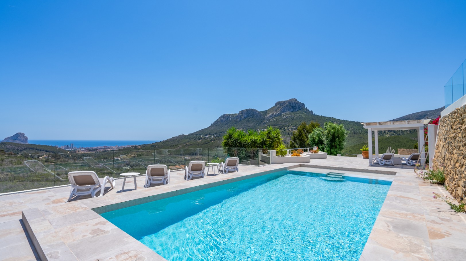Rectangular outdoor pool featuring a natural stone terrace, glass railings, and panoramic views of the Mediterranean coast and mountain ranges.