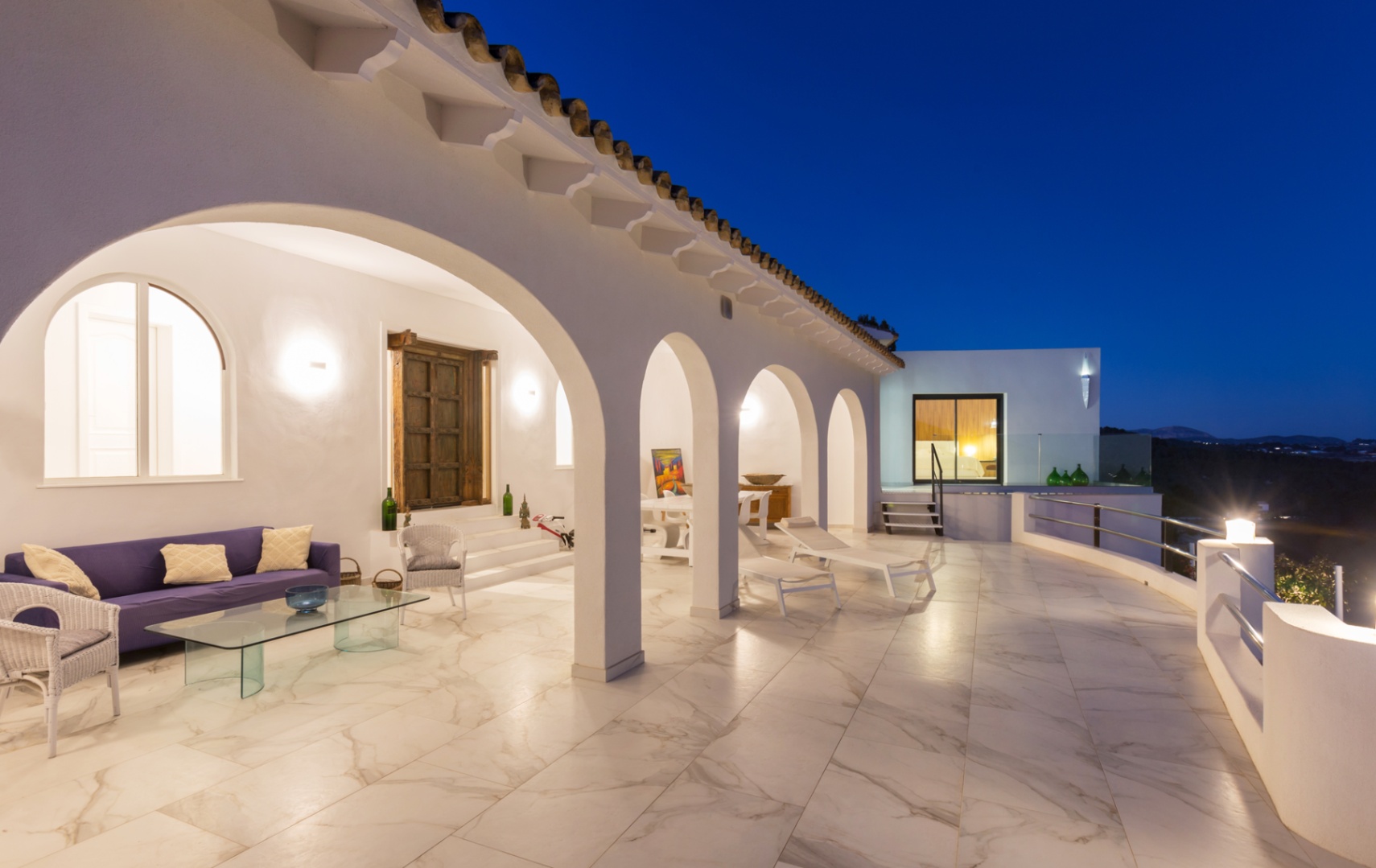 Spacious covered terrace at twilight featuring white arches, marble-effect flooring, a purple lounge area, and sun loungers overlooking the valley.