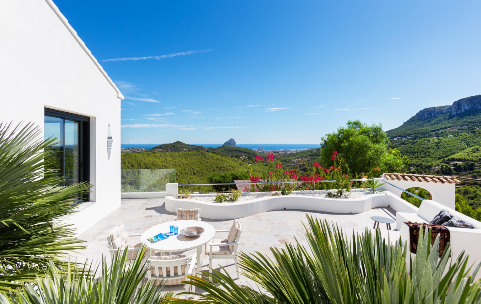 Spacious stone-paved terrace with a white dining set and curved planters, offering panoramic views of the Mediterranean coastline and Peñón de Ifach.