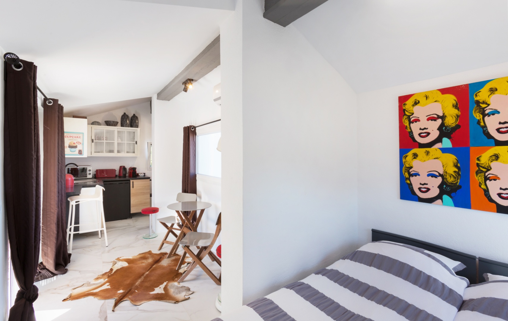 Studio space with pop-art decor, exposed dark beams, and marble-effect flooring. Features a compact kitchenette with red appliances and a dining area with a cowhide rug.