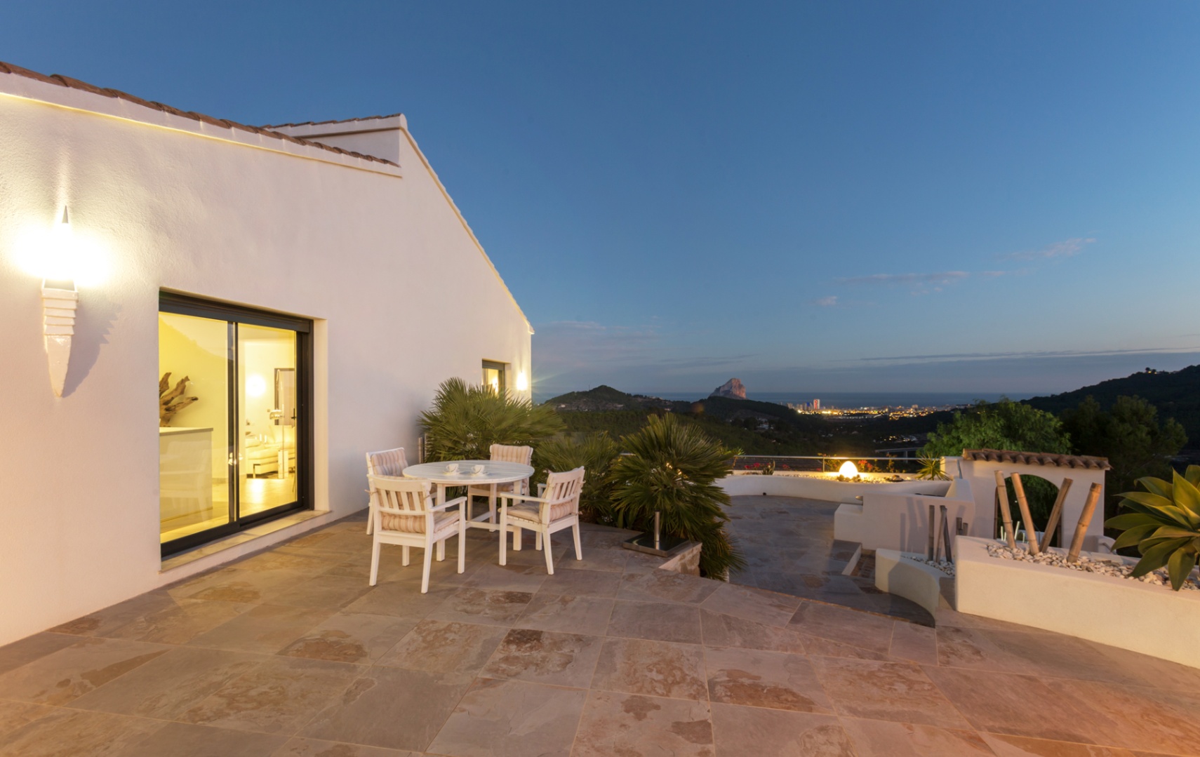 Terrace with natural stone flooring, white wooden dining set, and panoramic views of the Calpe coastline and Peñón de Ifach at dusk.
