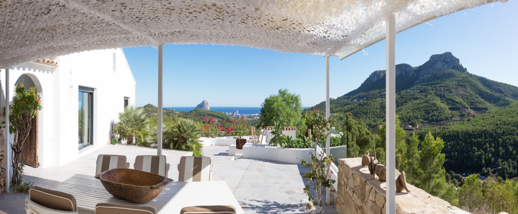 Terrace with white pergola and camouflage shading, featuring a dining area with views of the Mediterranean, the Peñón de Ifach, and forested mountains.