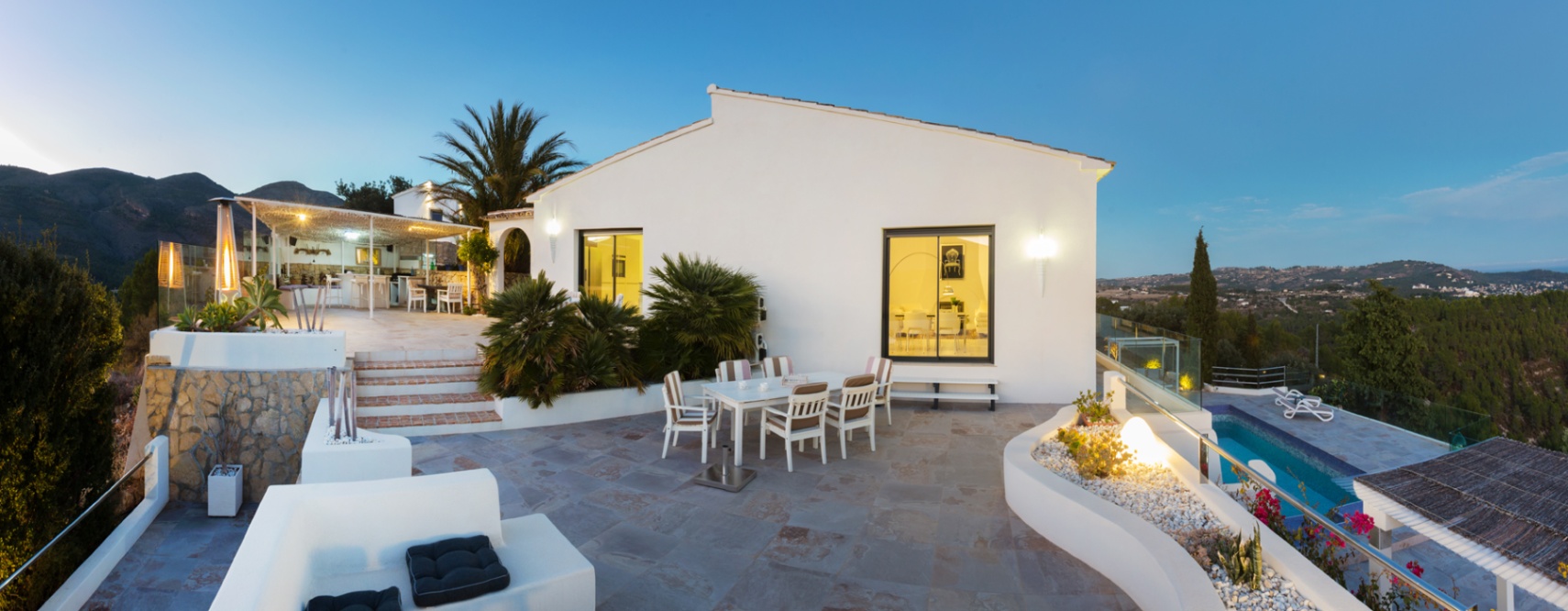 Wide stone terrace at dusk featuring a dining area, integrated planters, and panoramic valley views next to a white Mediterranean villa with illuminated interiors.