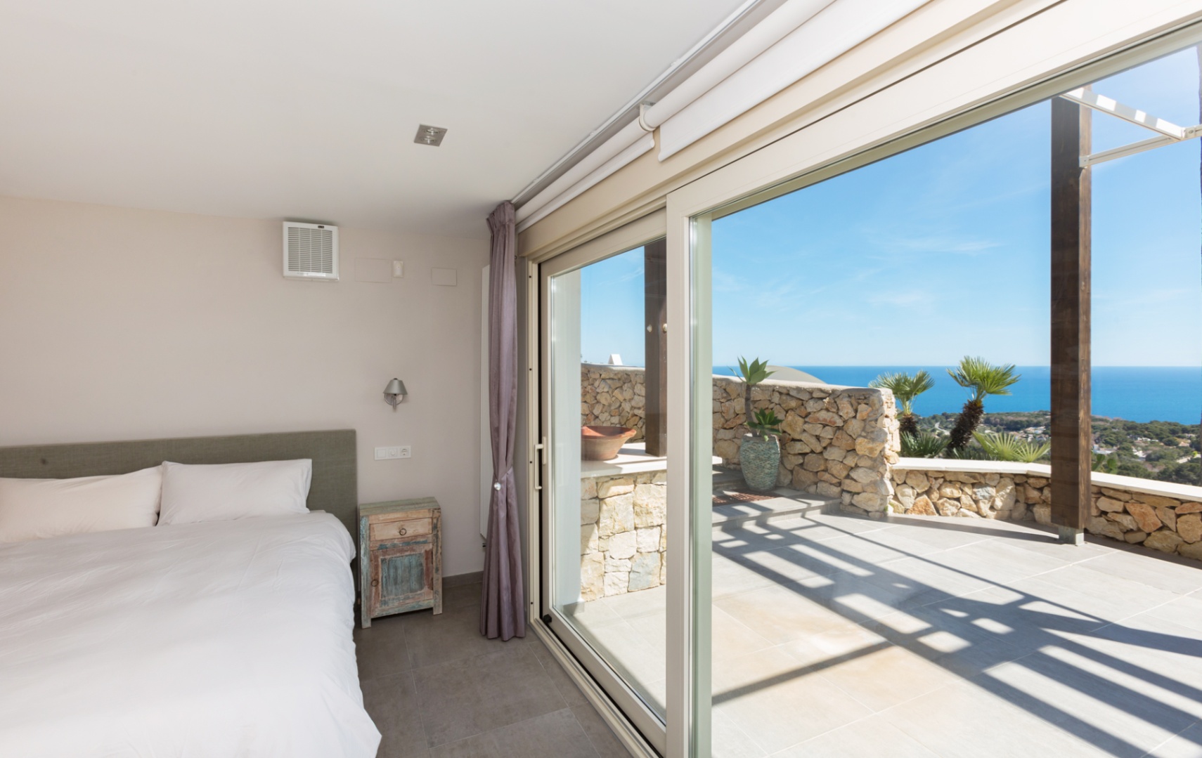 Bedroom with large sliding glass doors opening onto a stone-walled terrace with panoramic Mediterranean Sea views and rustic accents.