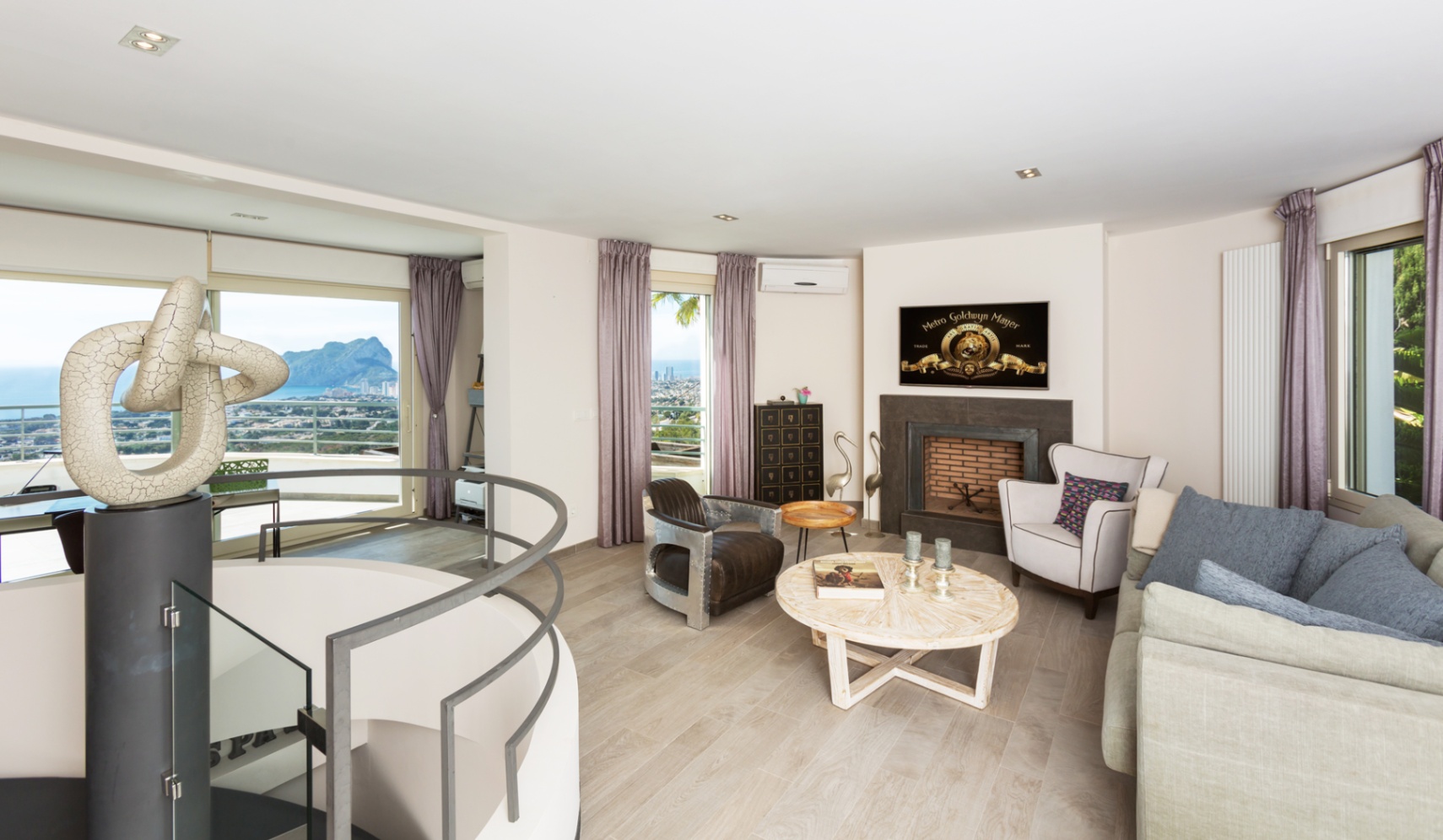 Contemporary living room featuring a spiral staircase with a modern sculpture, a fireplace, and large windows overlooking the sea and Peñón de Ifach.
